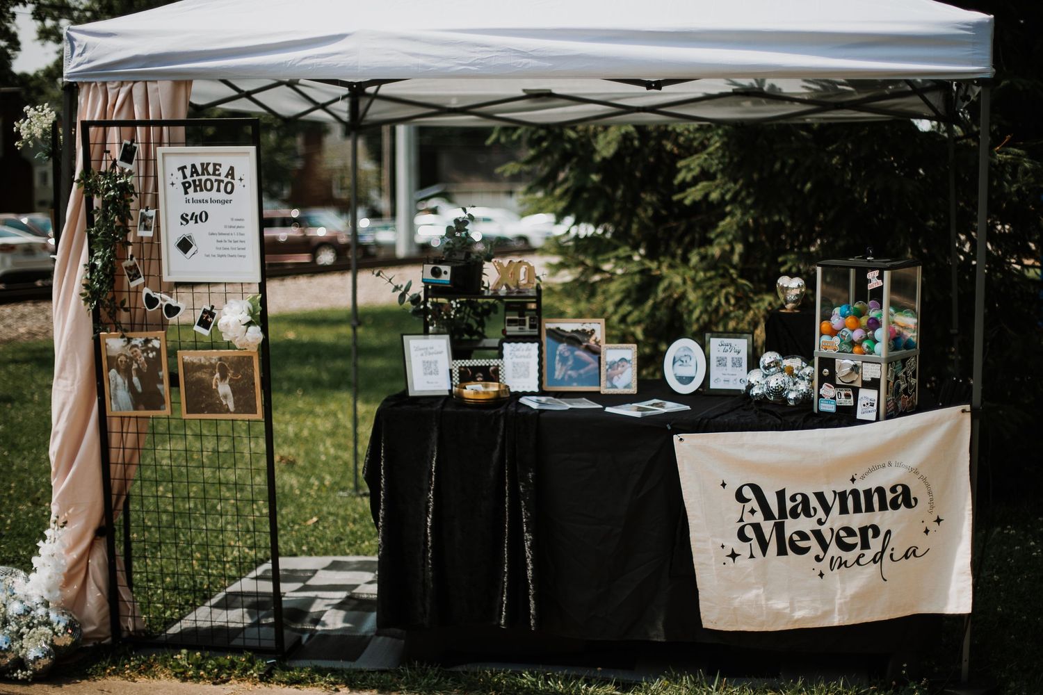 Outdoor photo booth setup under white canopy tent with black draped table and Maynina Meyer Media branded banner.