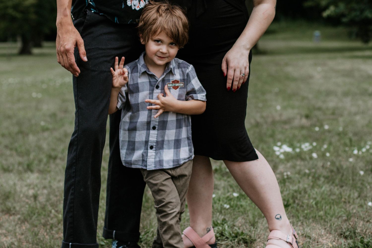 Cheerful young boy waving between parents legs on green grass in outdoor park setting in Dayton, Ohio