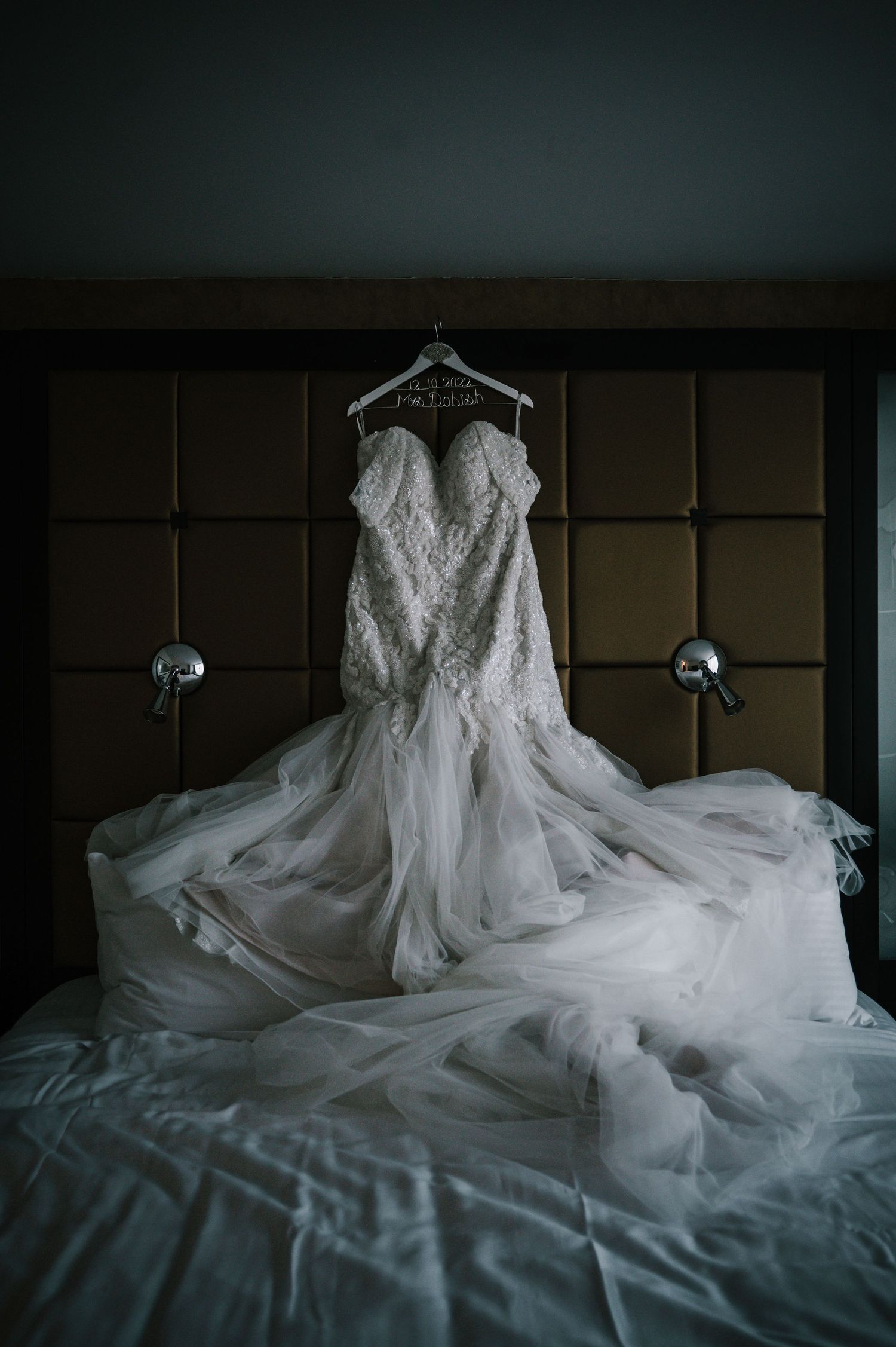 Elegant lace mermaid wedding dress hanging on personalized hanger over hotel bed.