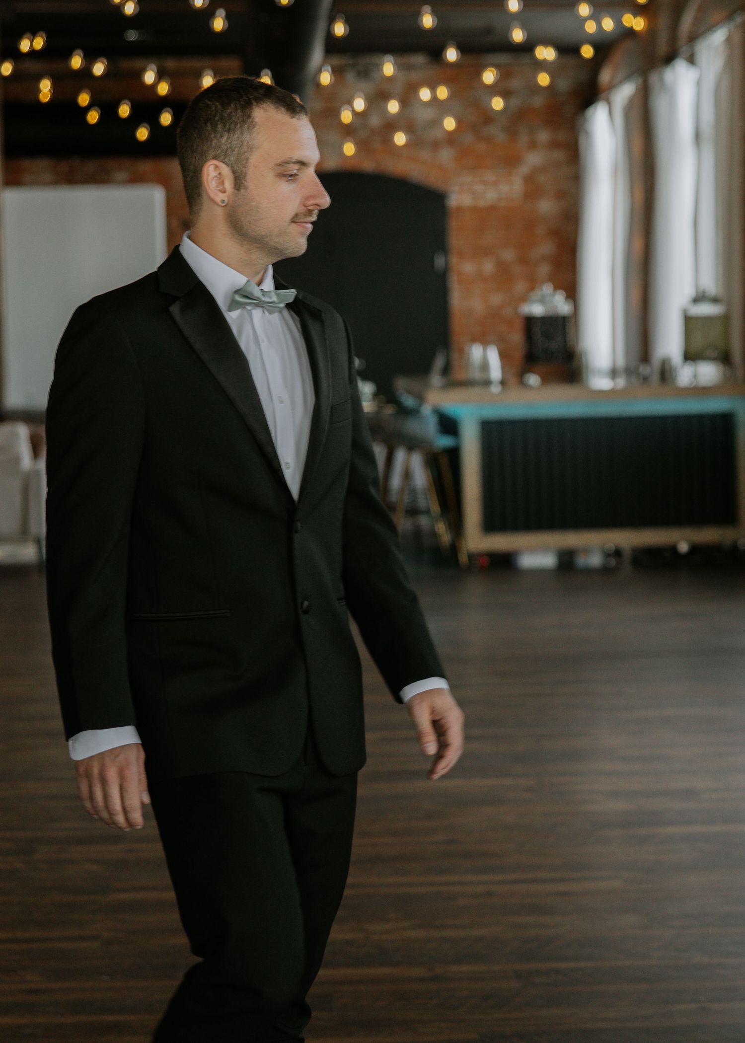 Groom in black tuxedo walks through elegant wedding venue with string lights and brick walls.