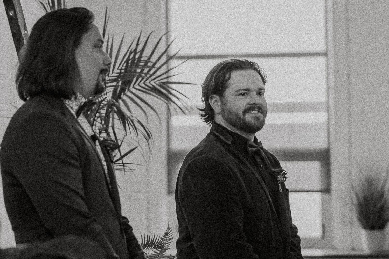 Two men in suits stand smiling at wedding ceremony in black and white photo.