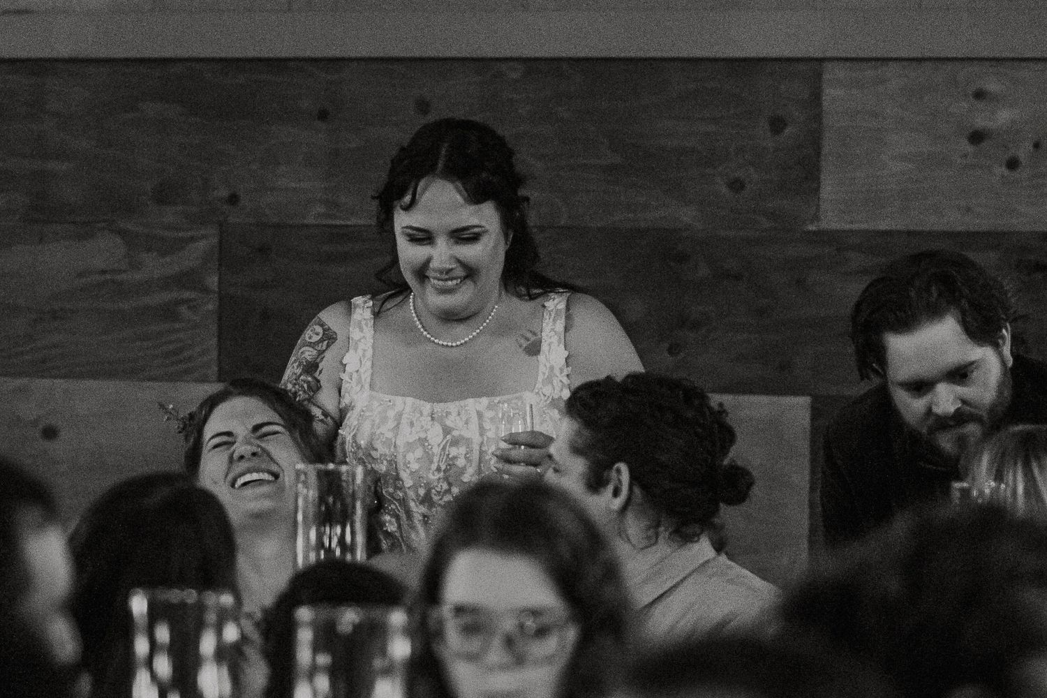 Black and white photo of a tattooed bride smiling warmly at laughing wedding reception guests.