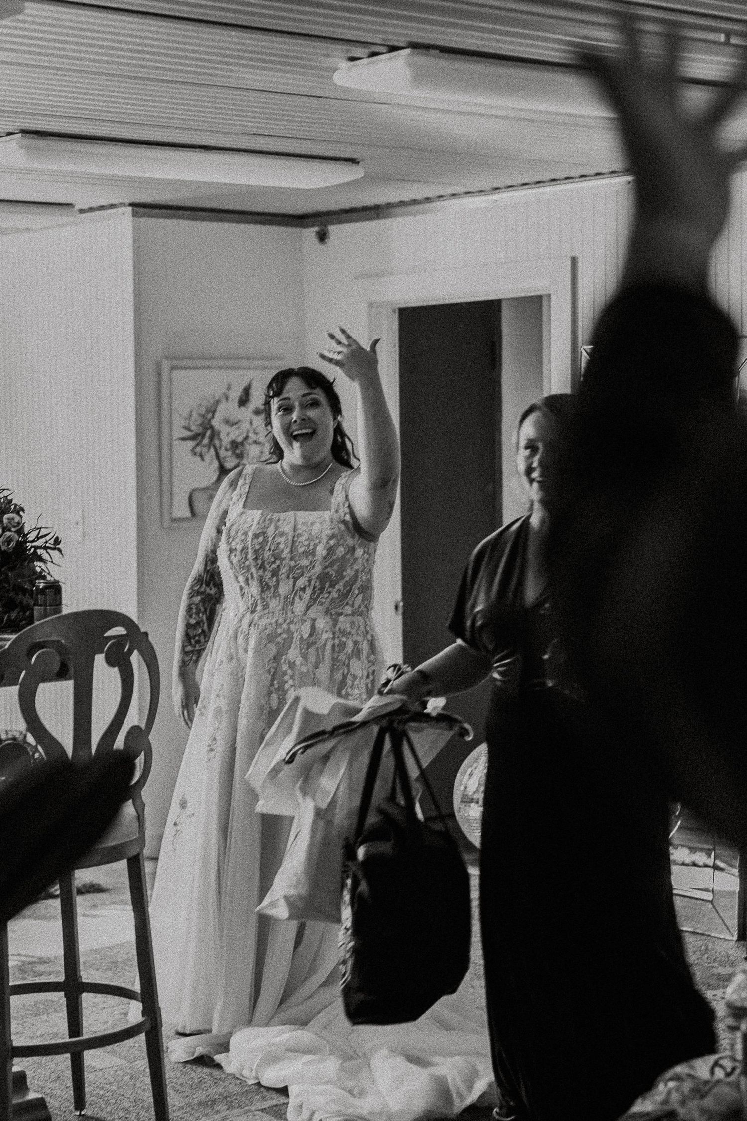 Black and white photo of joyful bride in lace gown waving hand with guest beside her.