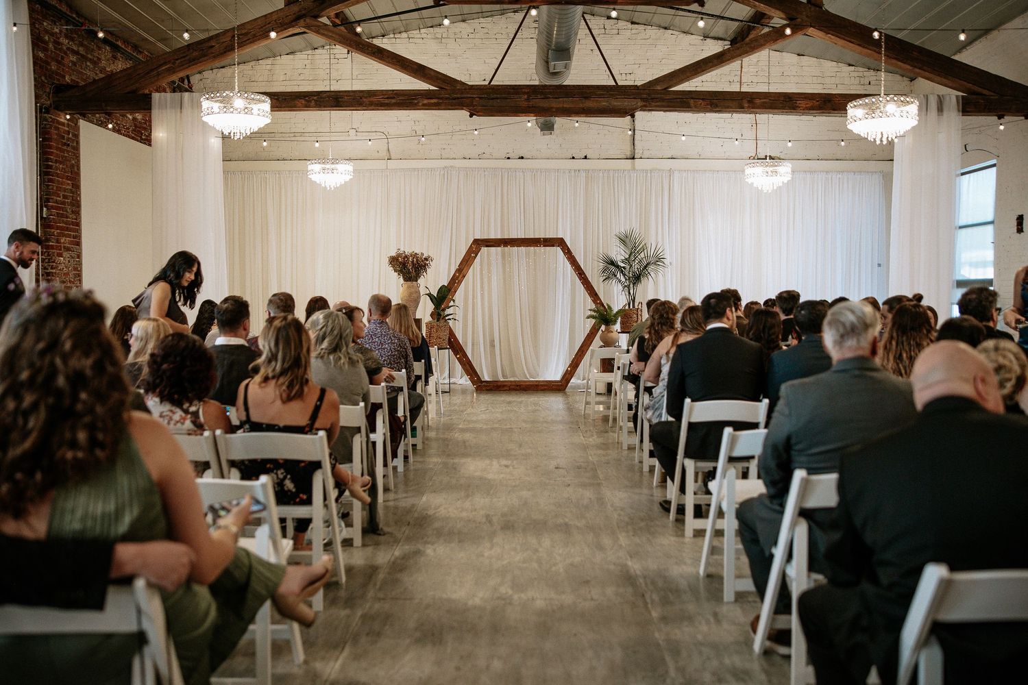 Wide shot of wedding ceremony with hexagonal wooden arch, white chairs, and guests in industrial venue.