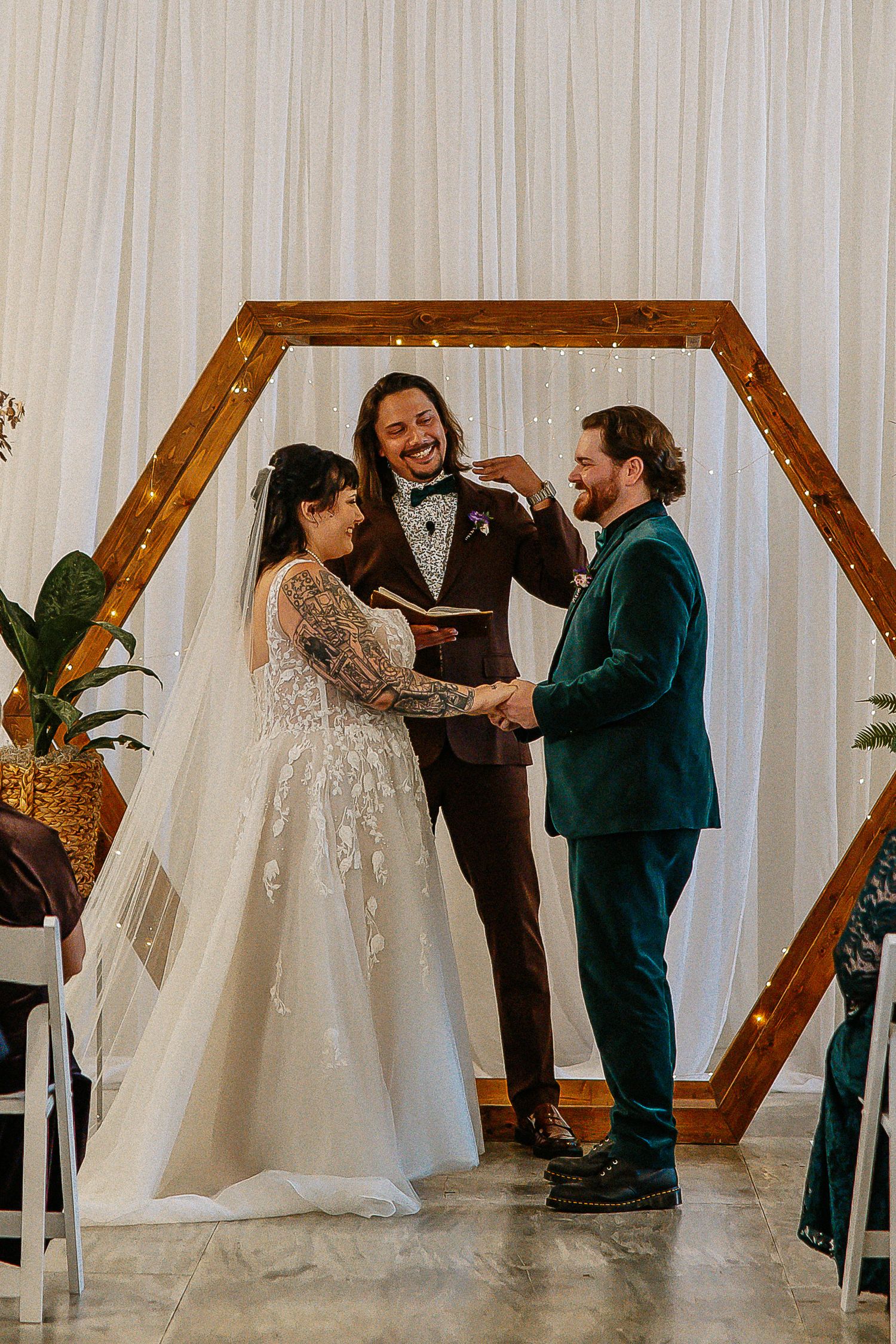 Bride, groom in green velvet suit, and officiant laughing joyfully during wedding ceremony.