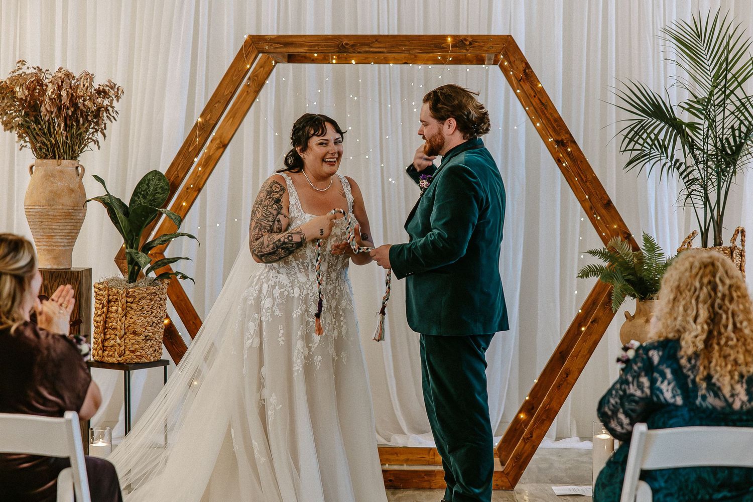 Laughing bride and groom in green velvet suit exchange rings at hexagonal wooden arch.