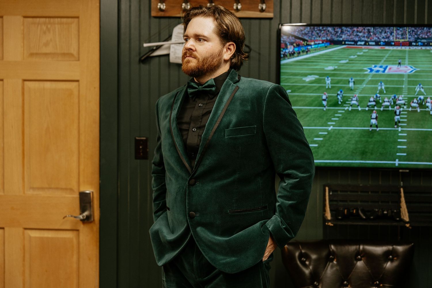 Bearded groom in green velvet suit with bow tie standing in dimly lit room with TV.