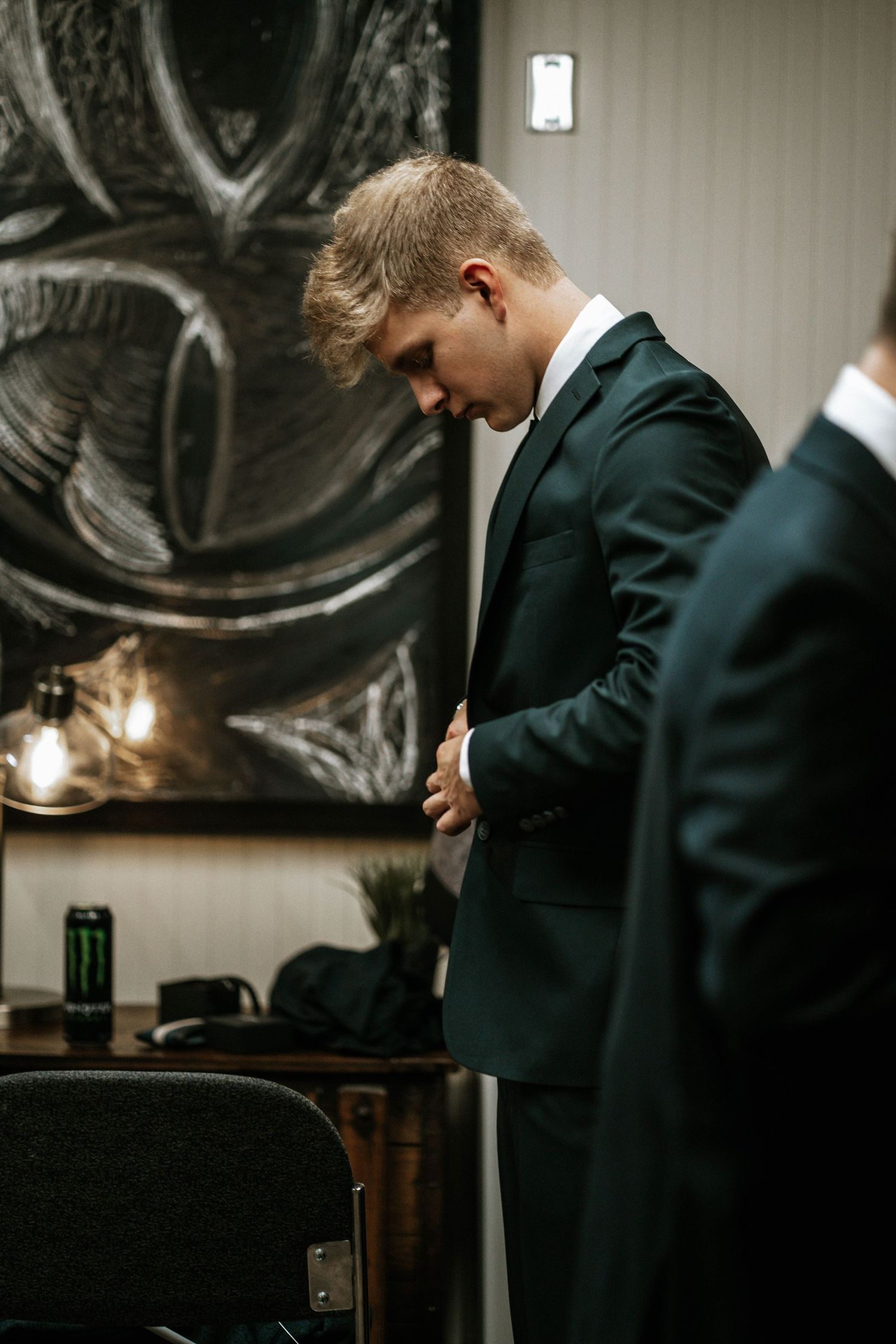 Young groomsman in dark green suit adjusting jacket in dimly lit room before wedding ceremony.