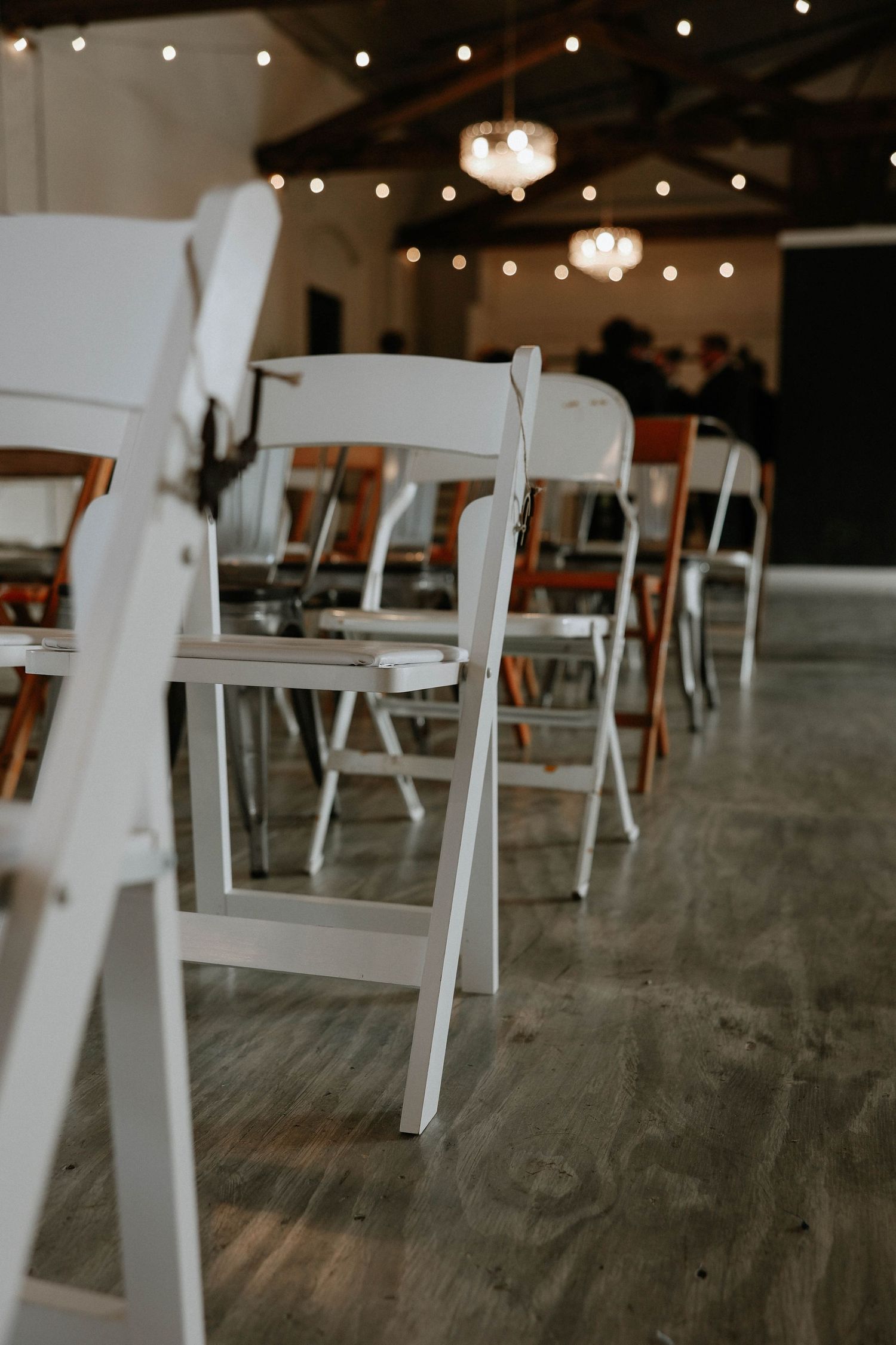 Rows of white folding chairs arranged on hardwood floors under string lights in an elegant event venue.
