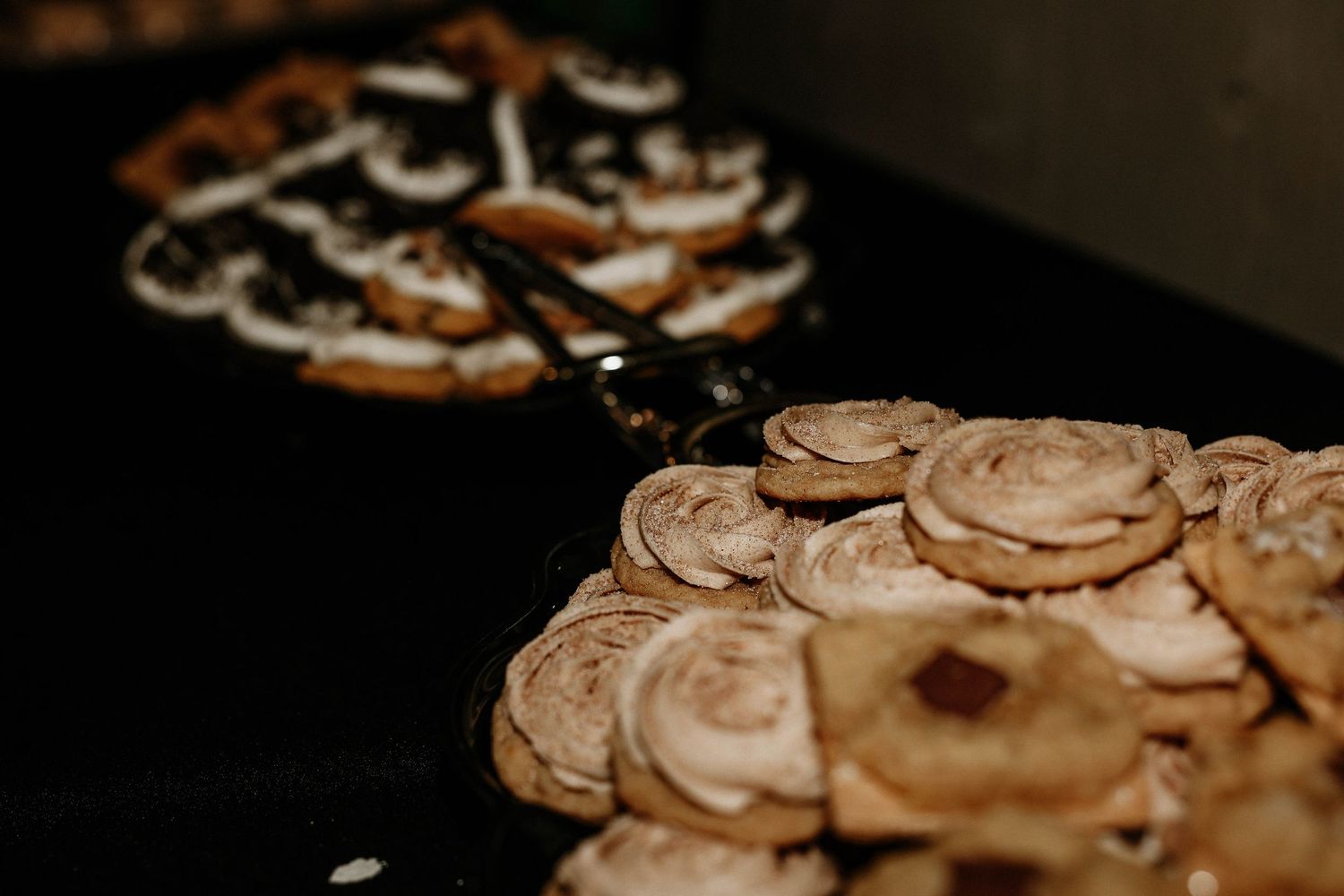 Assorted pastries including cinnamon rolls and cream-topped cookies displayed on a dark buffet table.