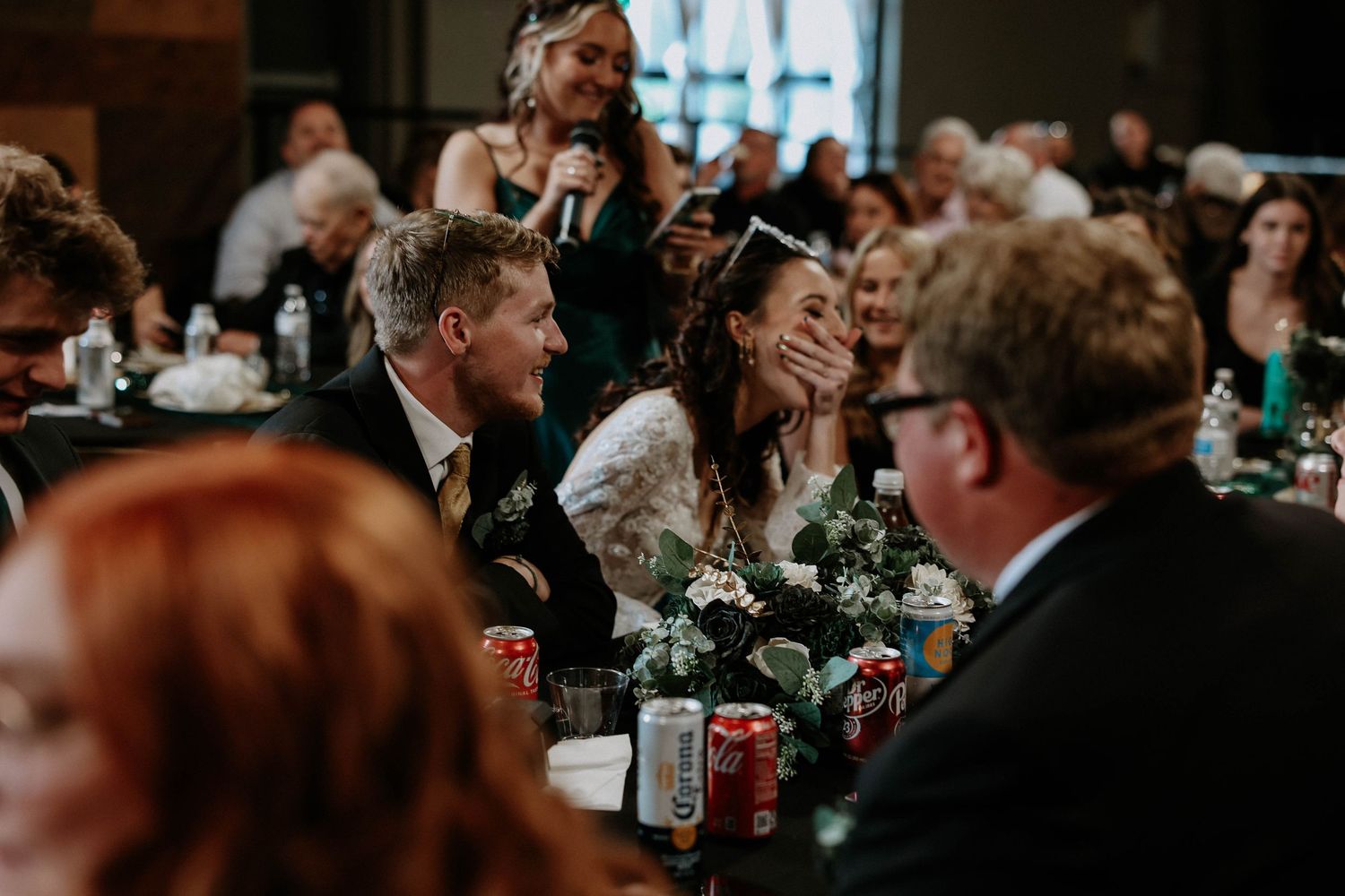 Bride and groom laughing joyfully at wedding reception dinner table surrounded by guests in elegant venue.
