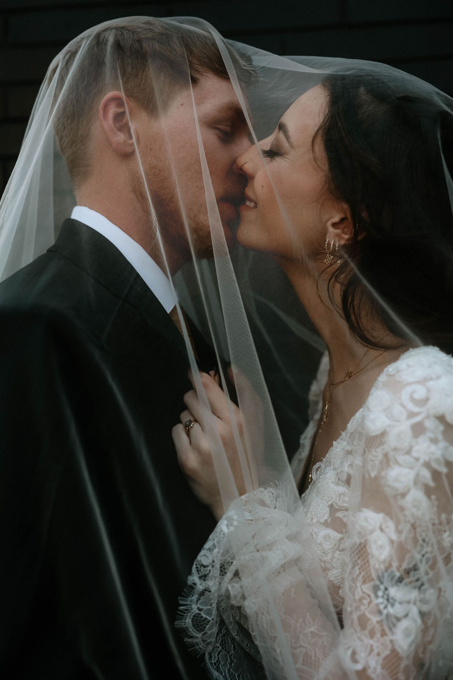 Bride and groom sharing a romantic kiss under a sheer wedding veil, wearing lace gown and black suit.