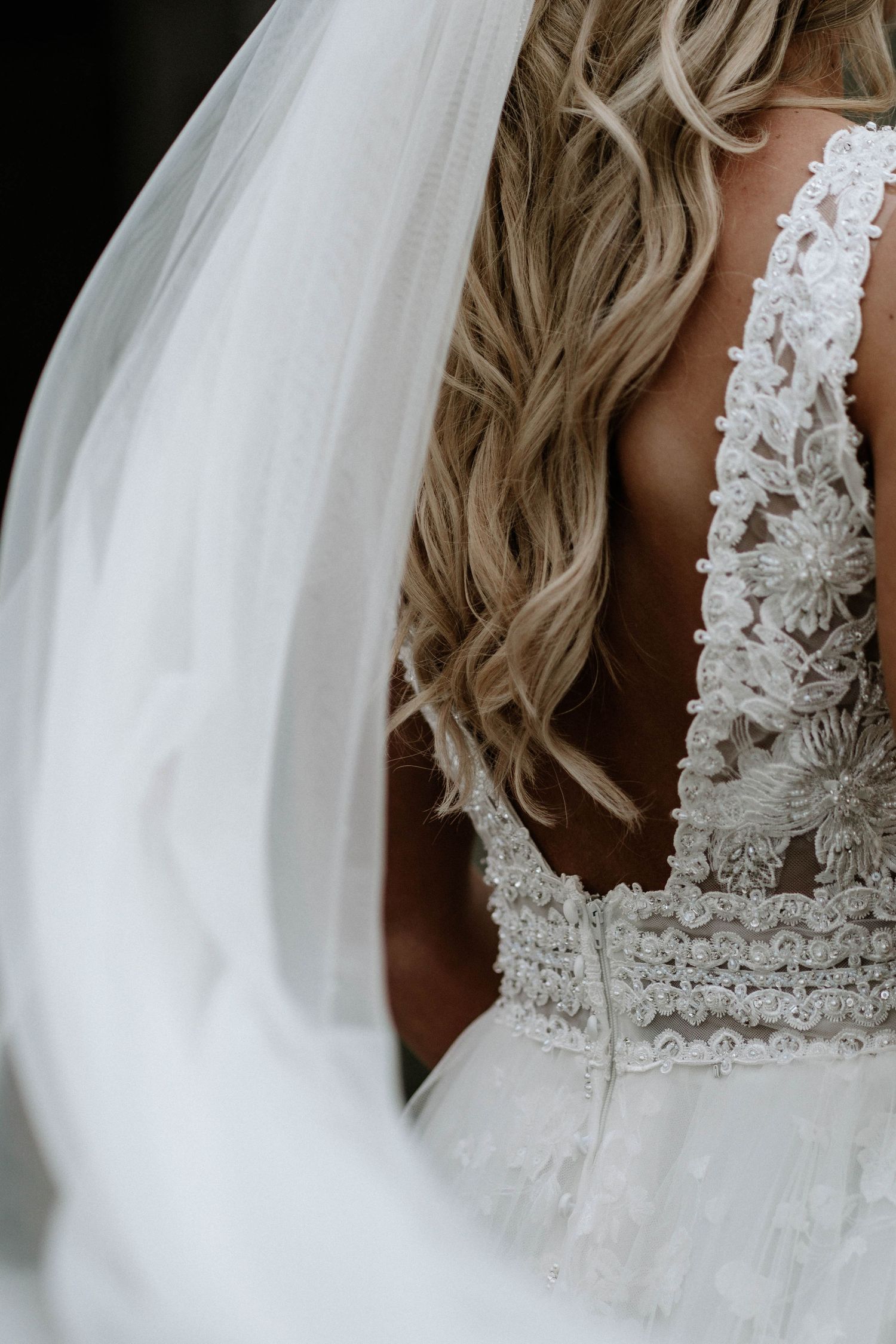 Close-up of bride's lace open-back wedding dress with flowing veil and curled blonde hair.