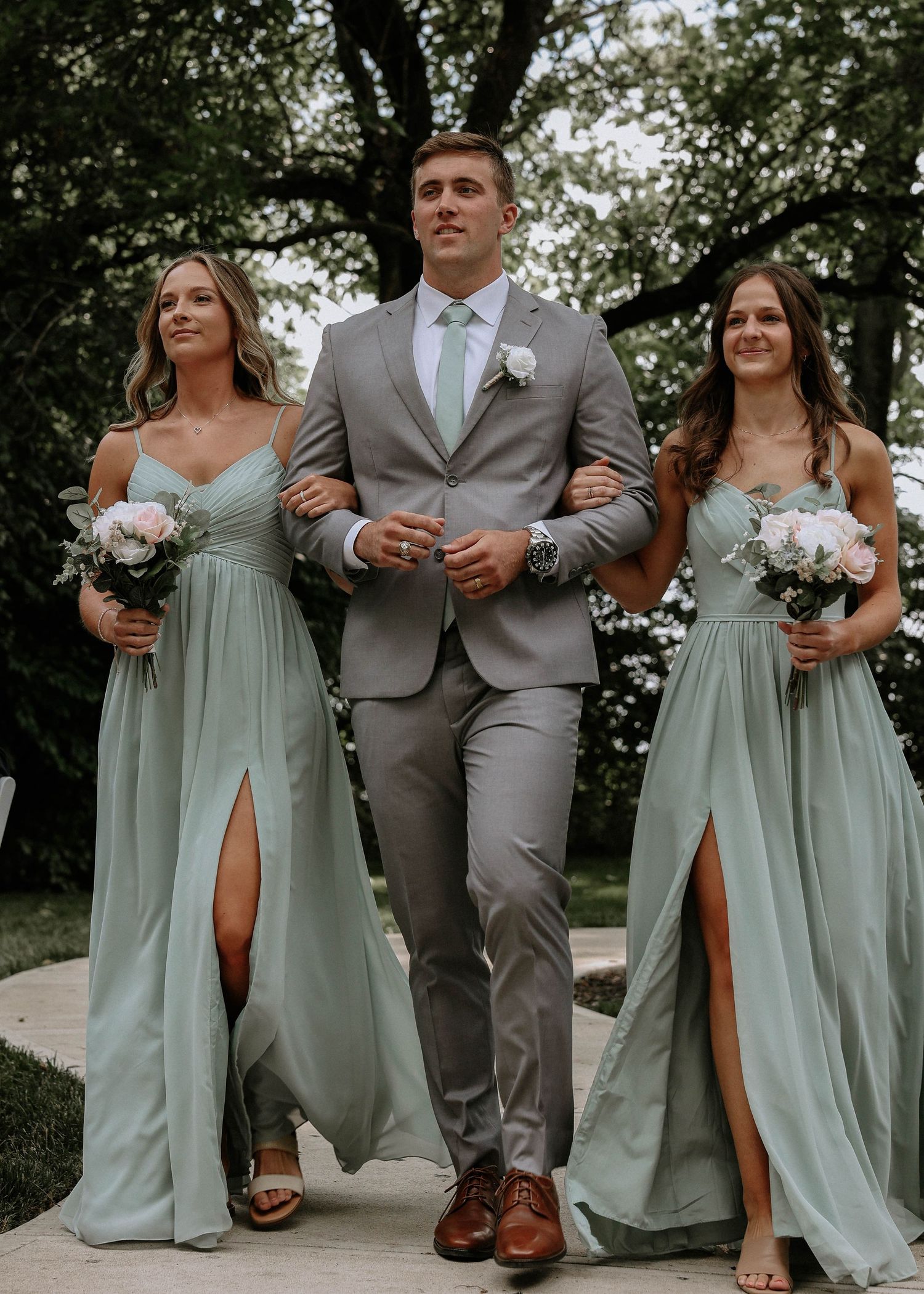 Groomsman in gray suit escorts two bridesmaids in sage green dresses down outdoor wedding aisle.