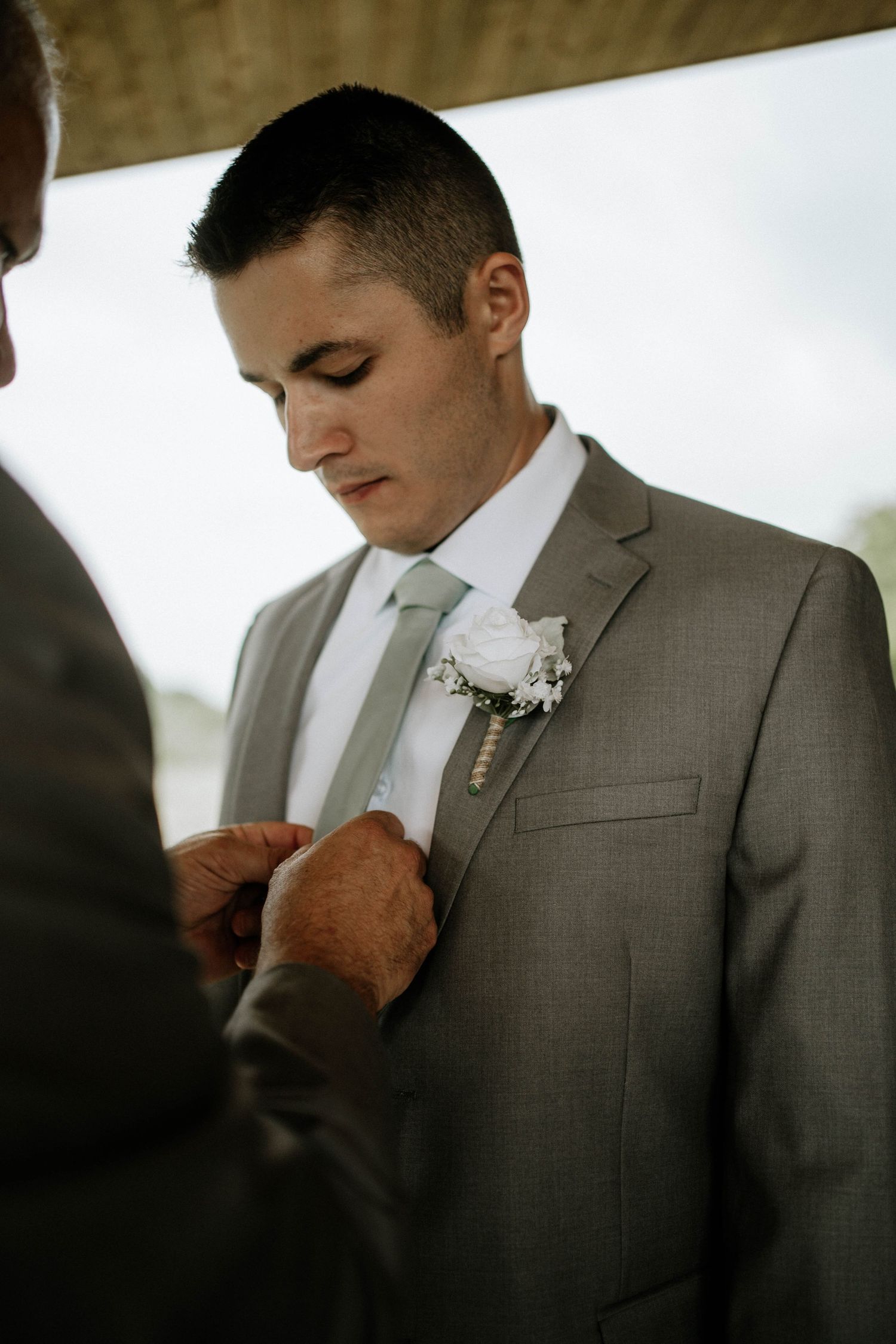 Groom in a gray suit having his white rose boutonniere pinned on before the wedding.