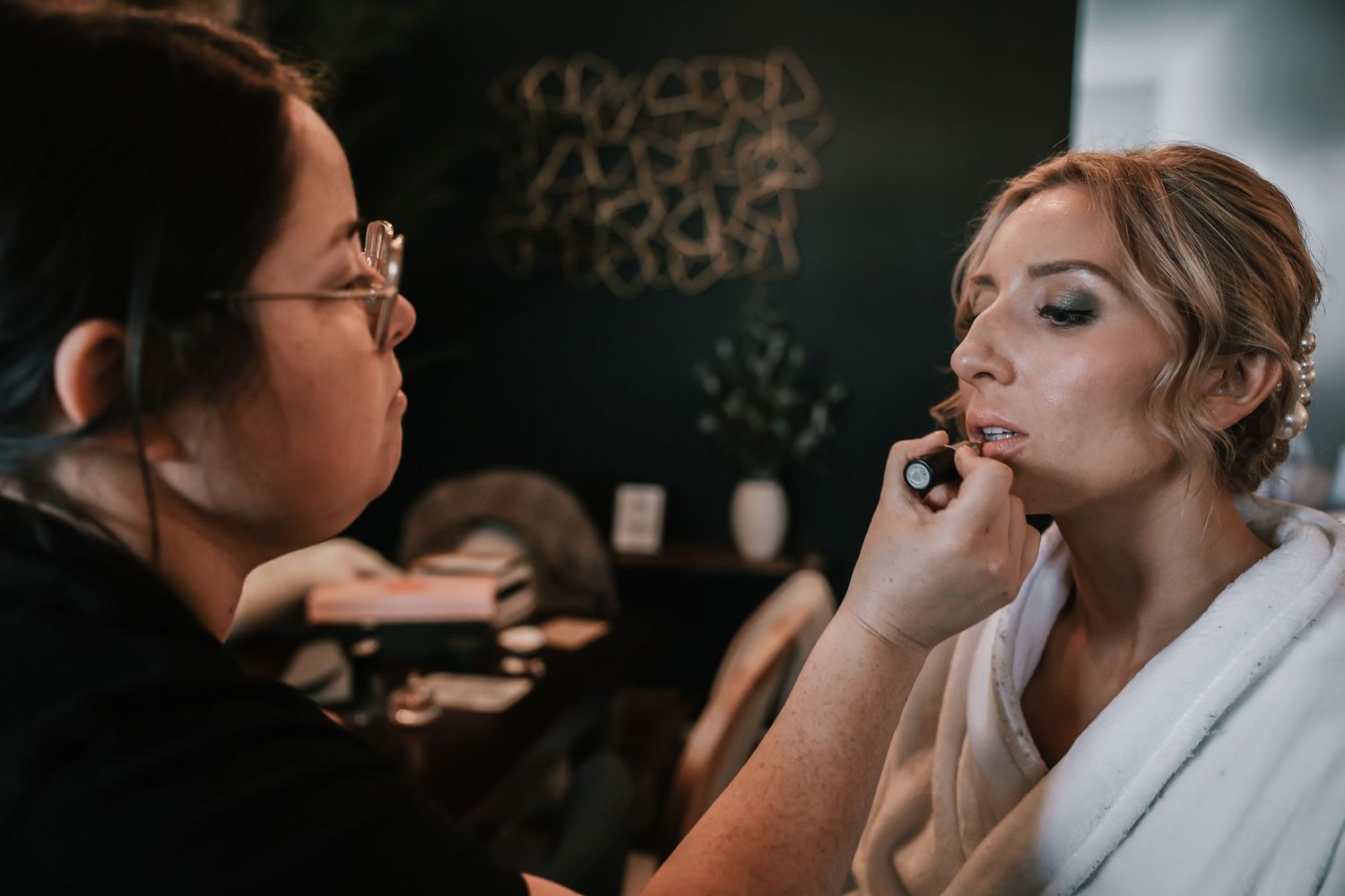 Professional makeup artist applying lipstick to a bride in a white robe before her wedding ceremony.