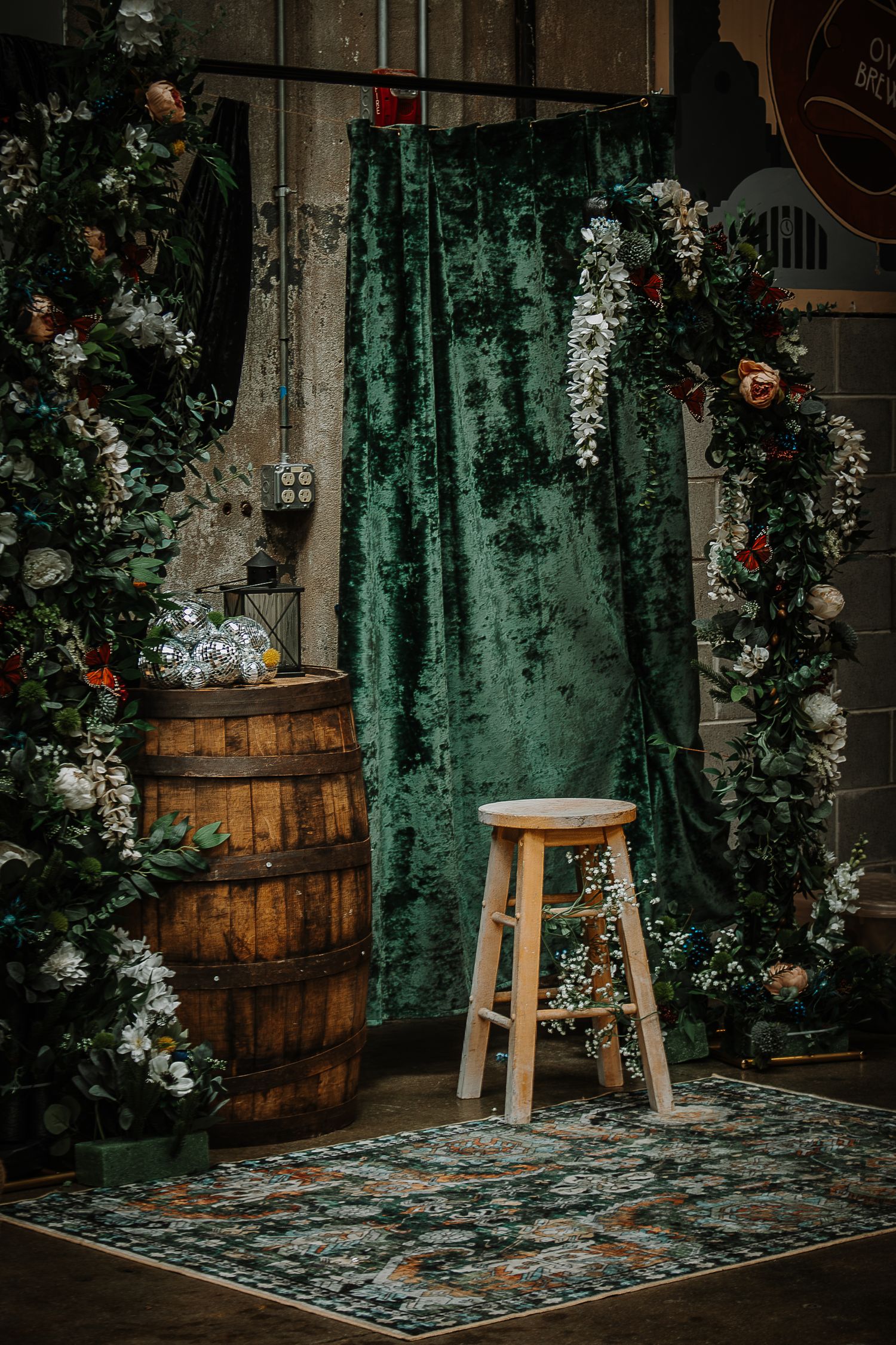 Rustic wedding photo booth with green velvet drape, wooden barrel, floral garlands, and wooden stool on vintage rug.