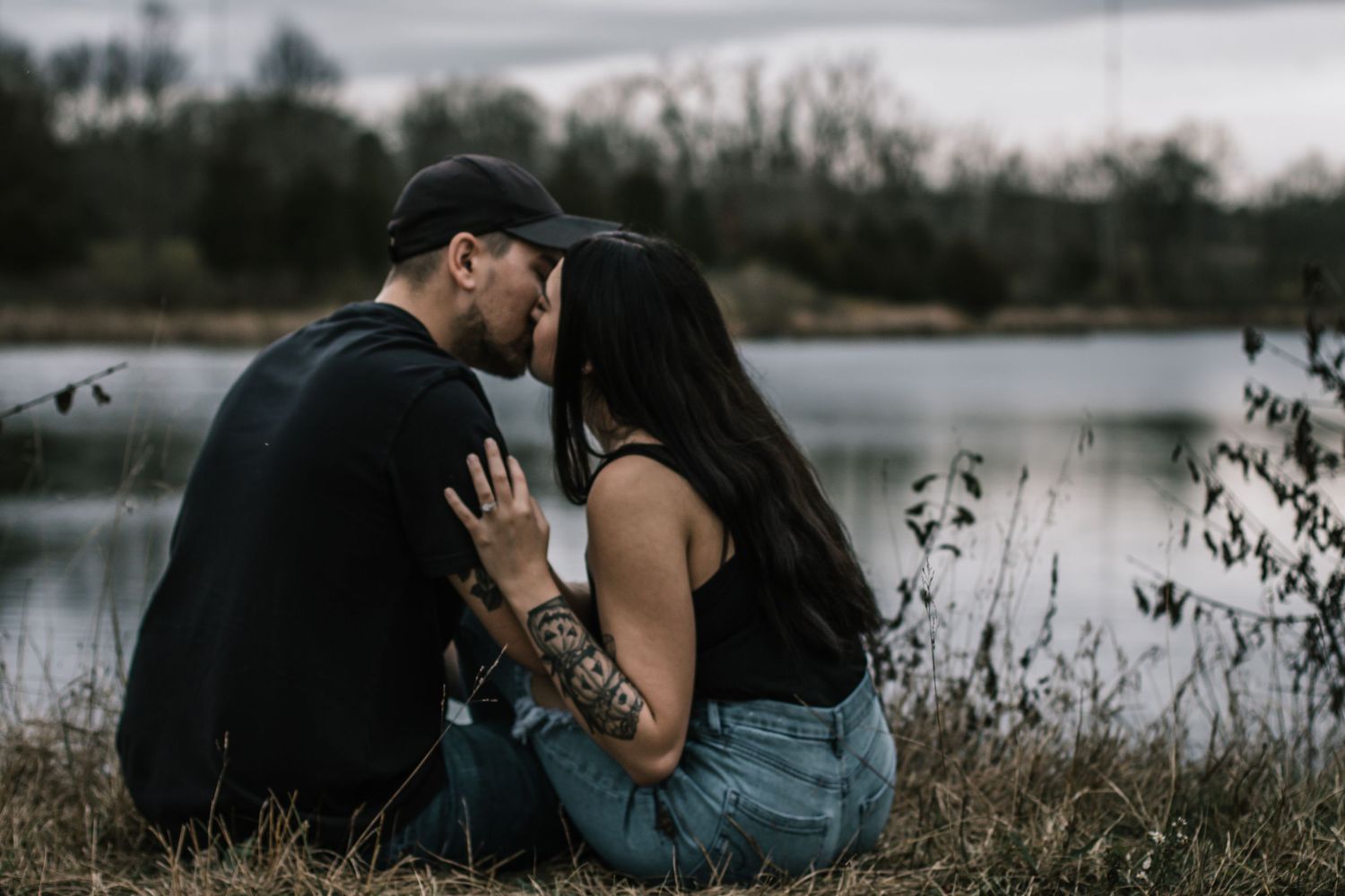 Couple by a lake at dusk, sharing a romantic moment with tattoos visible, surrounded by tall grass at Oakes Quarry Park in Dayton, Ohio