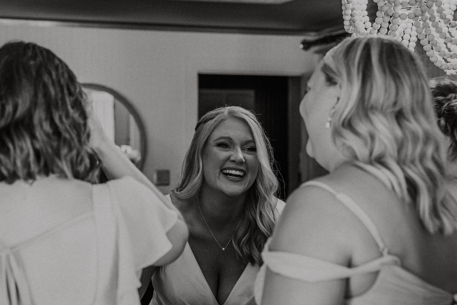 Black and white photo of a smiling bride laughing with bridesmaids in white dresses during wedding preparations.
