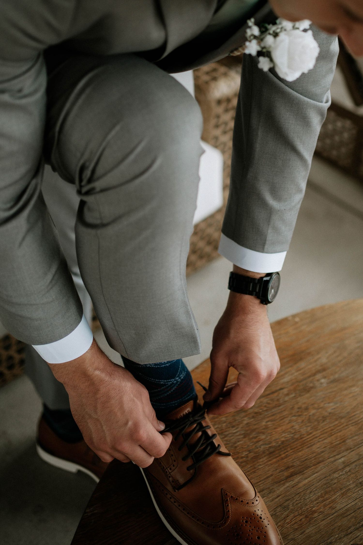 Groom in gray suit tying brown leather dress shoes with white boutonniere visible at wedding.