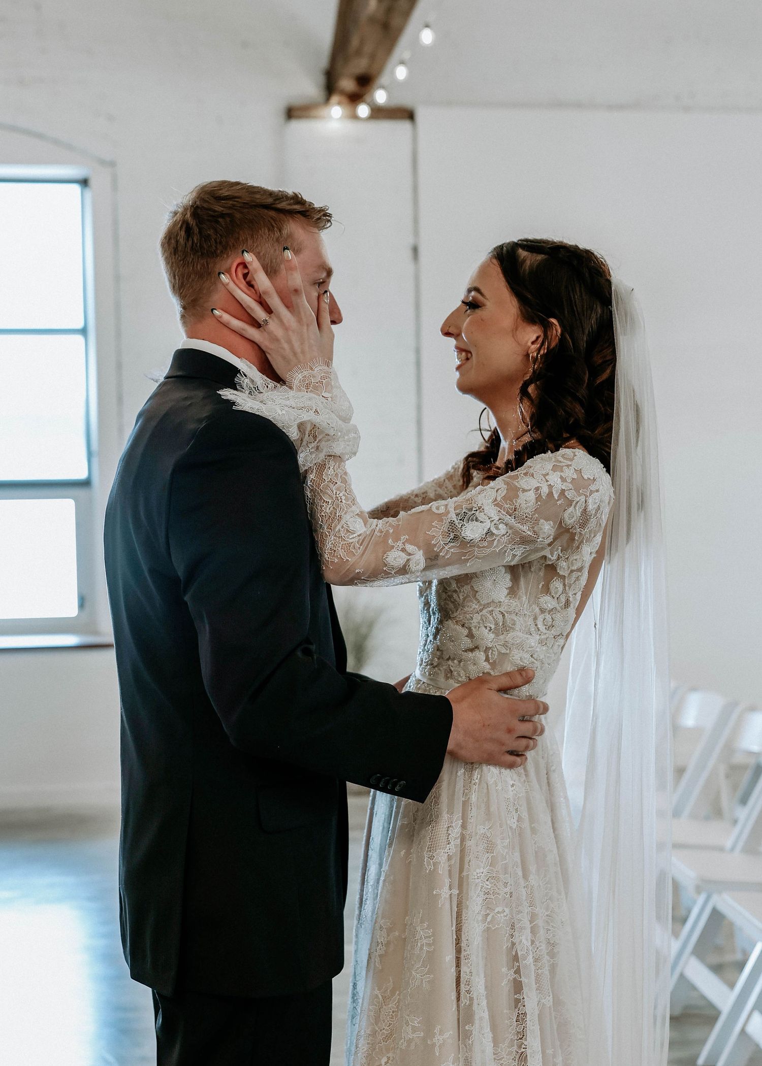 Bride in lace long-sleeve gown touches groom's face tenderly during romantic first look at indoor wedding venue.
