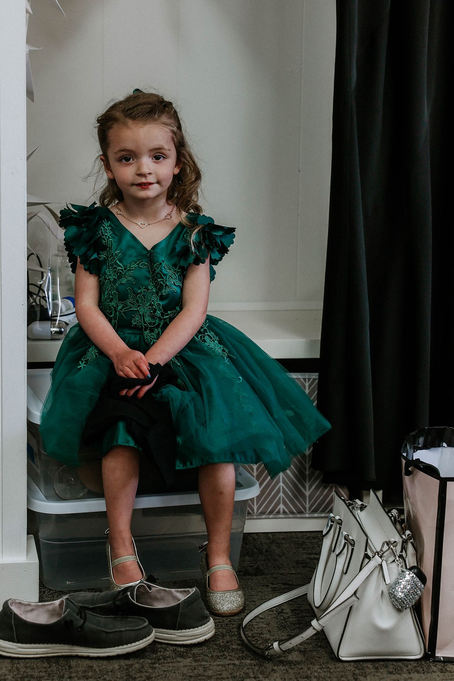 Young girl in emerald green tulle dress sits in dressing room, wearing silver shoes with handbag nearby.