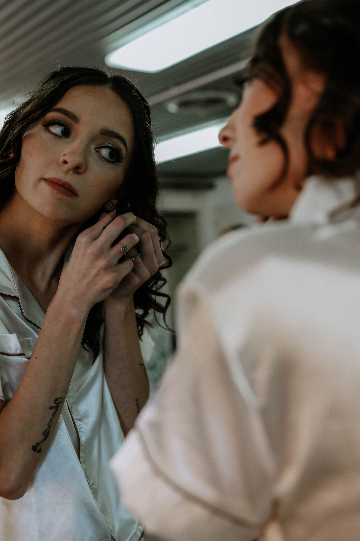 Bride putting on earrings while looking in mirror, wearing white satin robe before wedding ceremony.