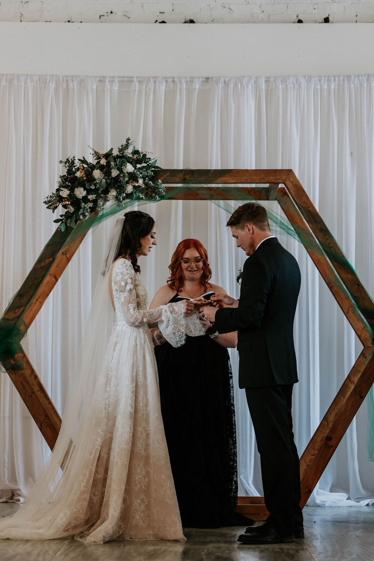 Couple exchanging rings under a hexagonal wooden arch with floral decor, officiated by a woman in black dress.