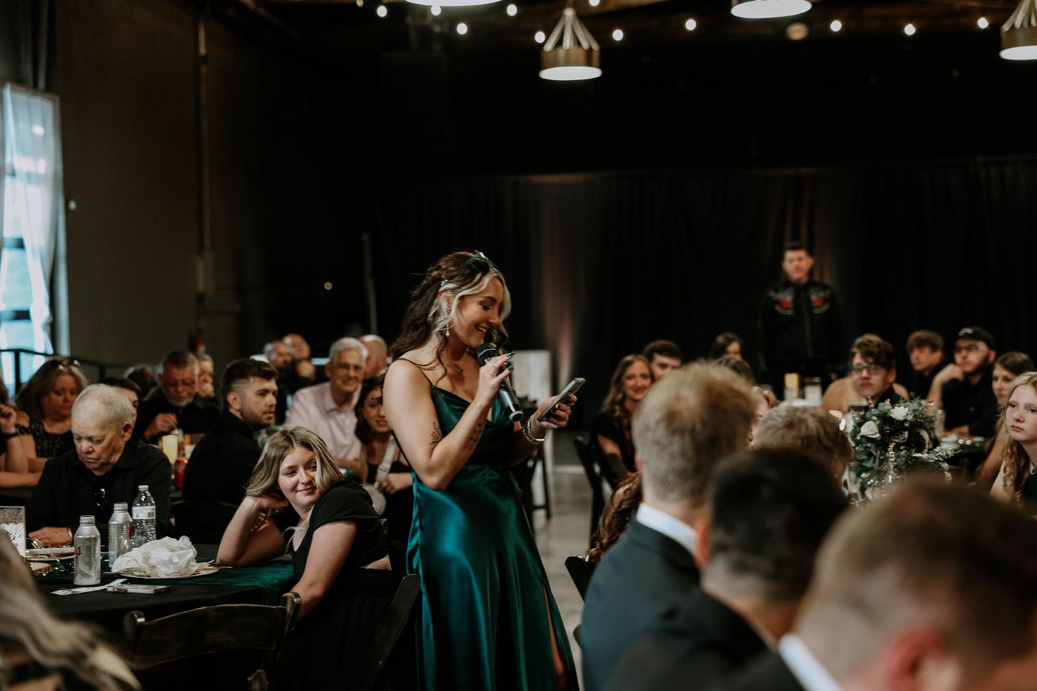 A woman in an emerald green gown gives a speech at an elegant wedding reception with seated guests.