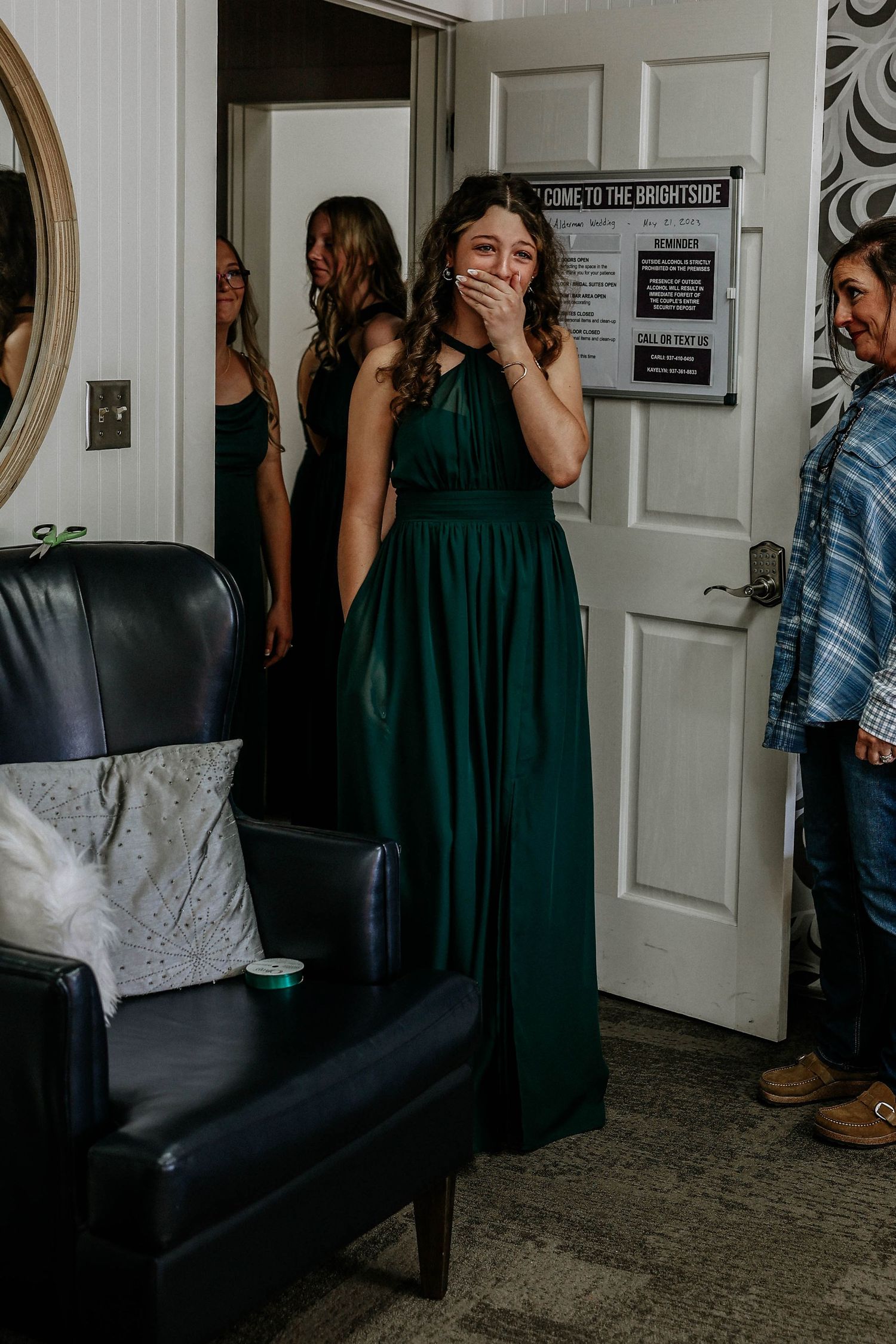 Bridesmaid in emerald green gown covering her mouth in surprise at a wedding venue.