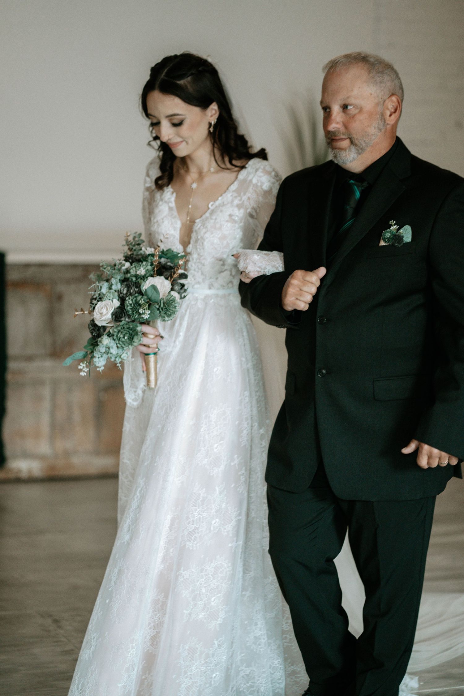 Bride in lace gown holding bouquet walks down aisle with older man in dark suit at elegant wedding ceremony.