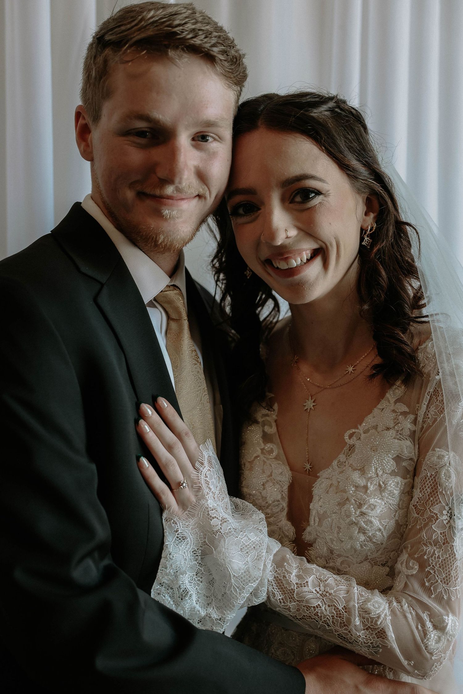 Smiling bride and groom pose together on their wedding day, bride wearing lace dress and veil, groom in dark suit with gold tie.