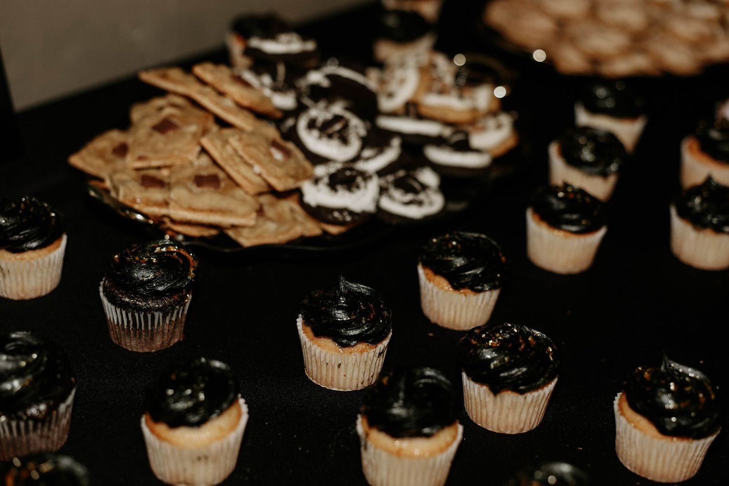 Dessert table with black frosted cupcakes, white cream cheese bites, and crackers on black tablecloth.