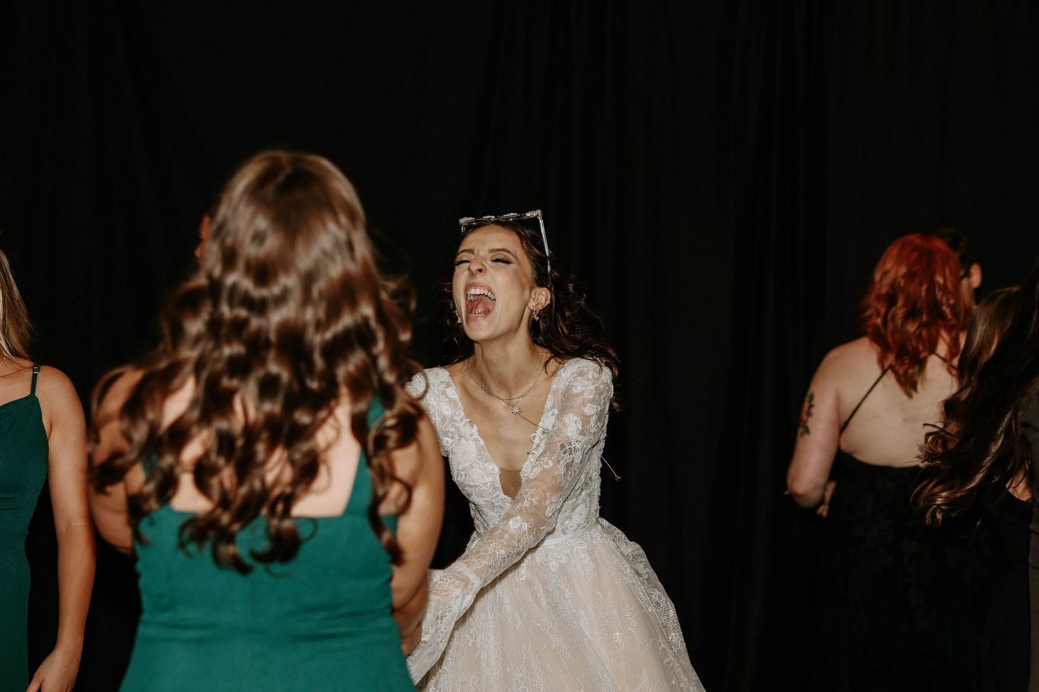 Bride in white lace gown laughing joyfully on dance floor surrounded by bridesmaids in emerald green dresses.