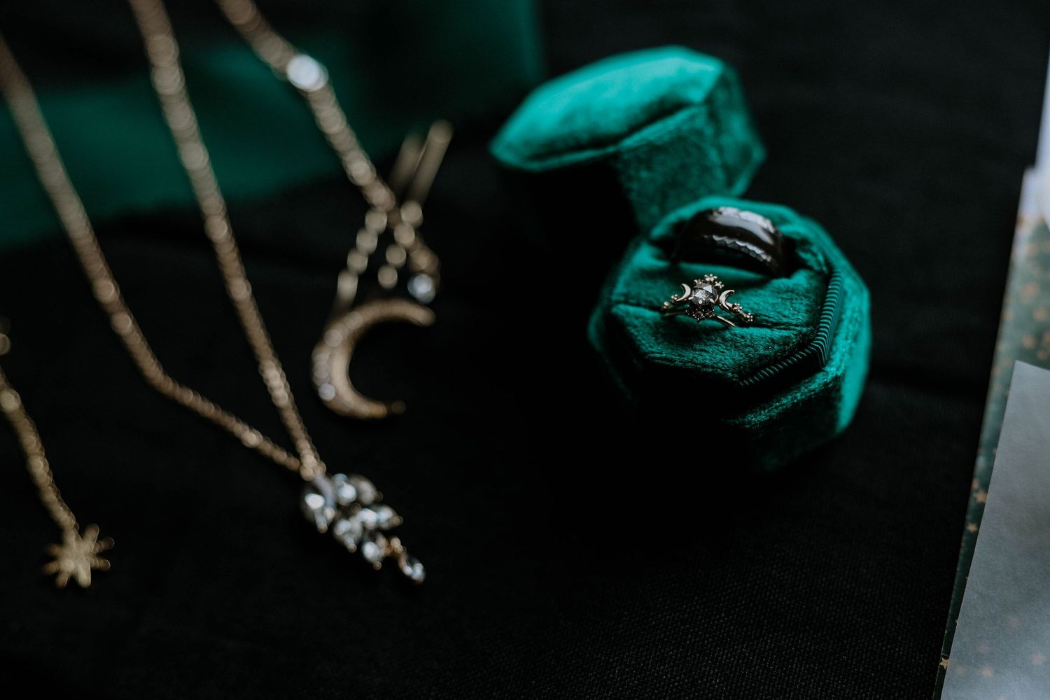 Wedding rings displayed in a green velvet ring box alongside delicate gold celestial necklaces on a dark surface.