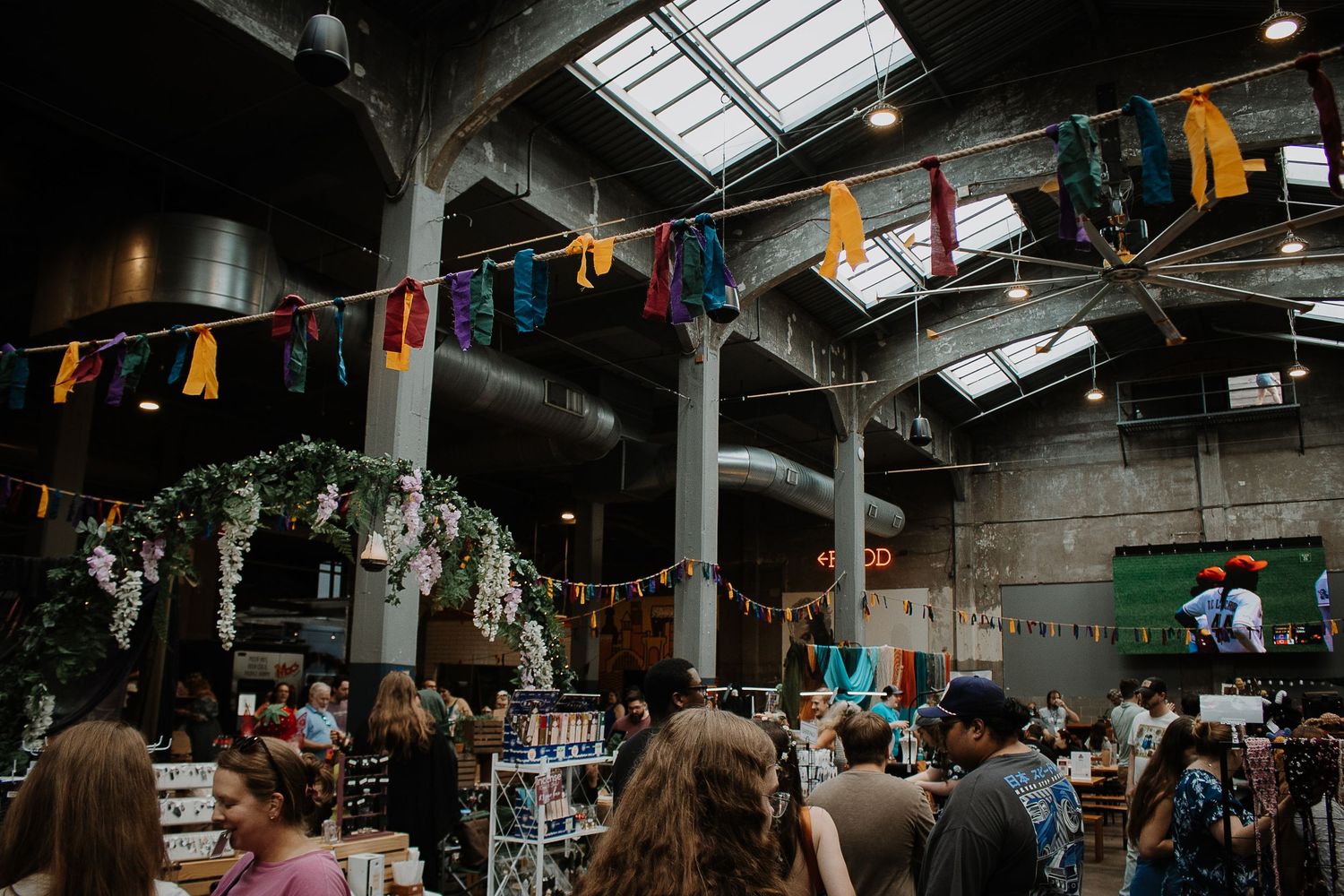 Colorful indoor market with bunting, floral arch, and crowds browsing vendor stalls in an industrial warehouse venue.