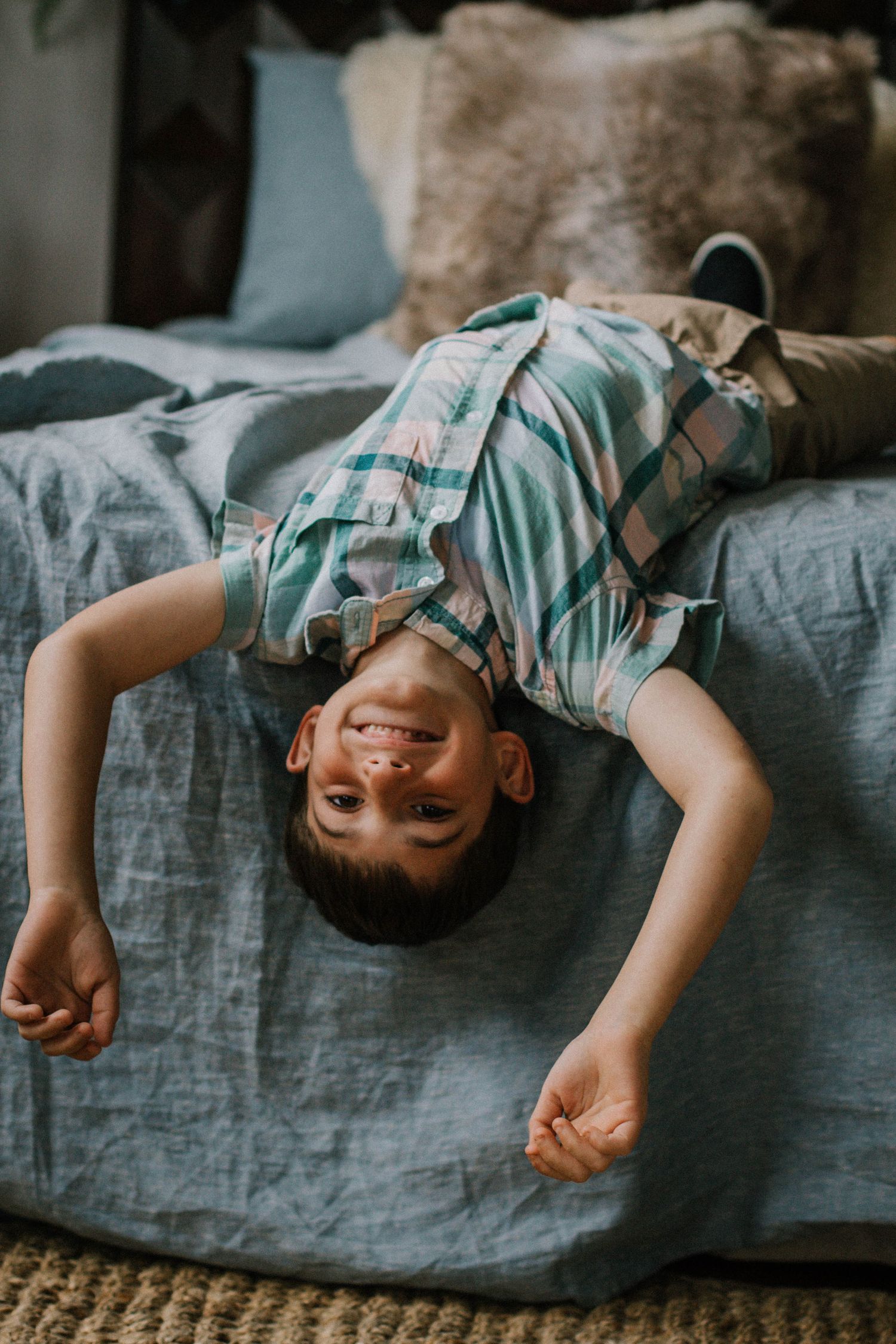 Smiling young boy hanging upside down off edge of bed in plaid shirt indoors at Mill Street Studios in Cincinnati, Ohio.