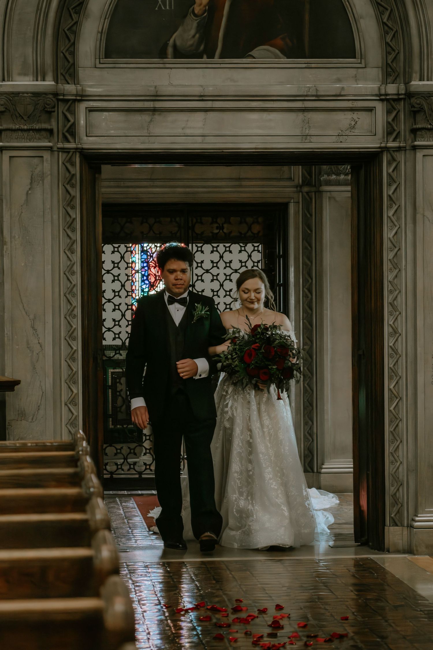 Bride in lace gown holding red bouquet walks down church aisle escorted by man in tuxedo in Cincinnati, Ohio