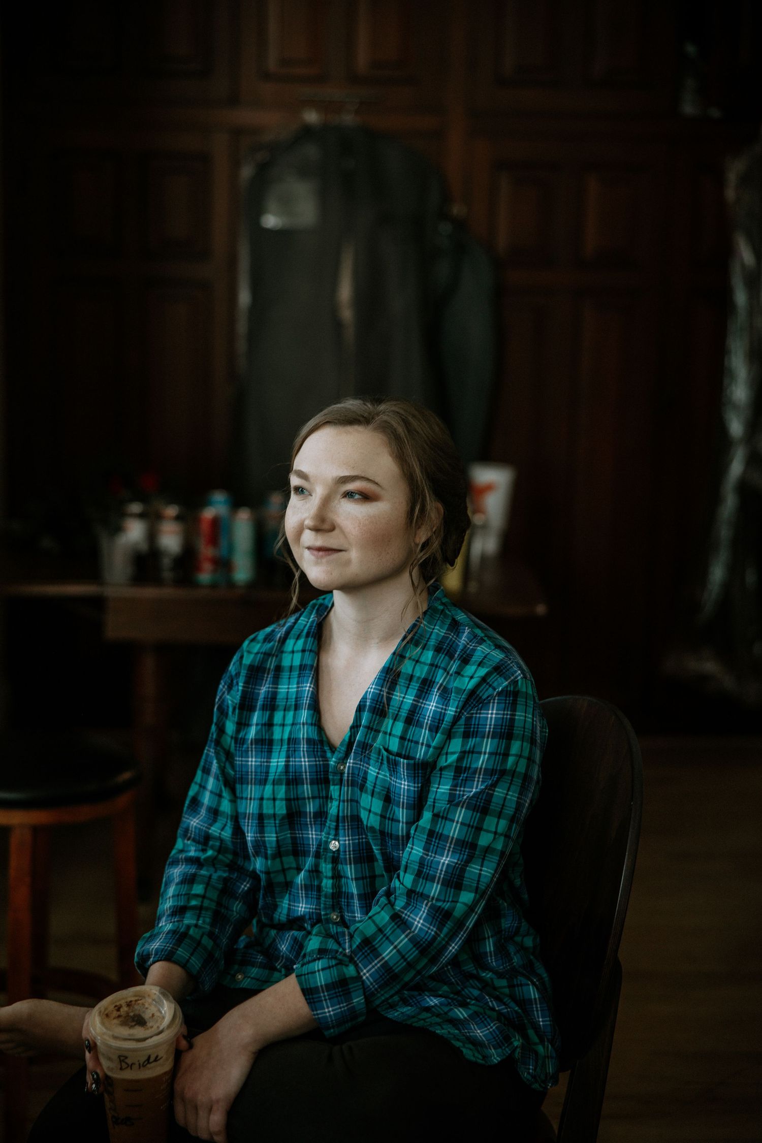 Smiling bridesmaid in green plaid shirt holding an iced drink while sitting in a dark wood room at Urban Artifact in Cincinnati, Ohio