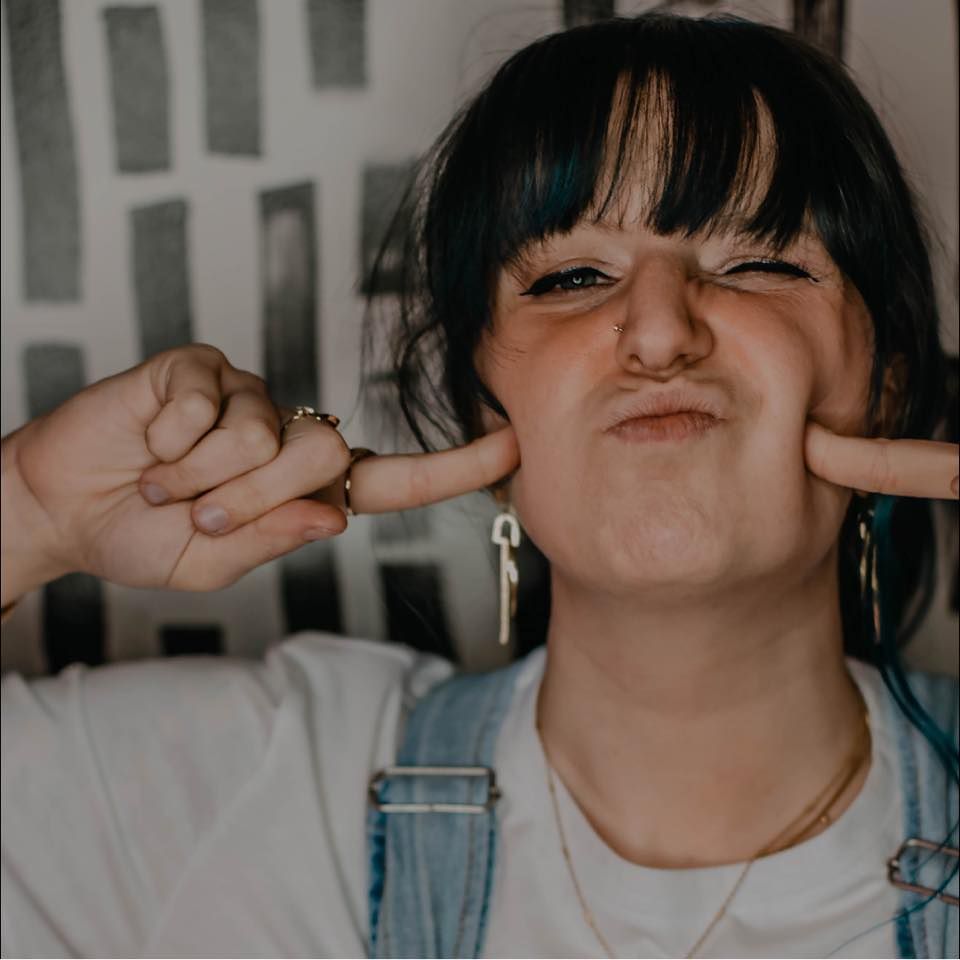 Alaynna Meyer Media making a funny face, squishing her cheeks with both hands, wearing overalls and a white tee.
