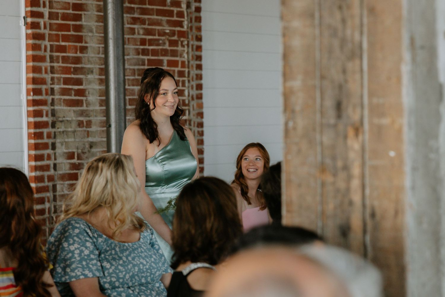 Woman in green satin dress stands smiling at indoor wedding reception with brick walls.