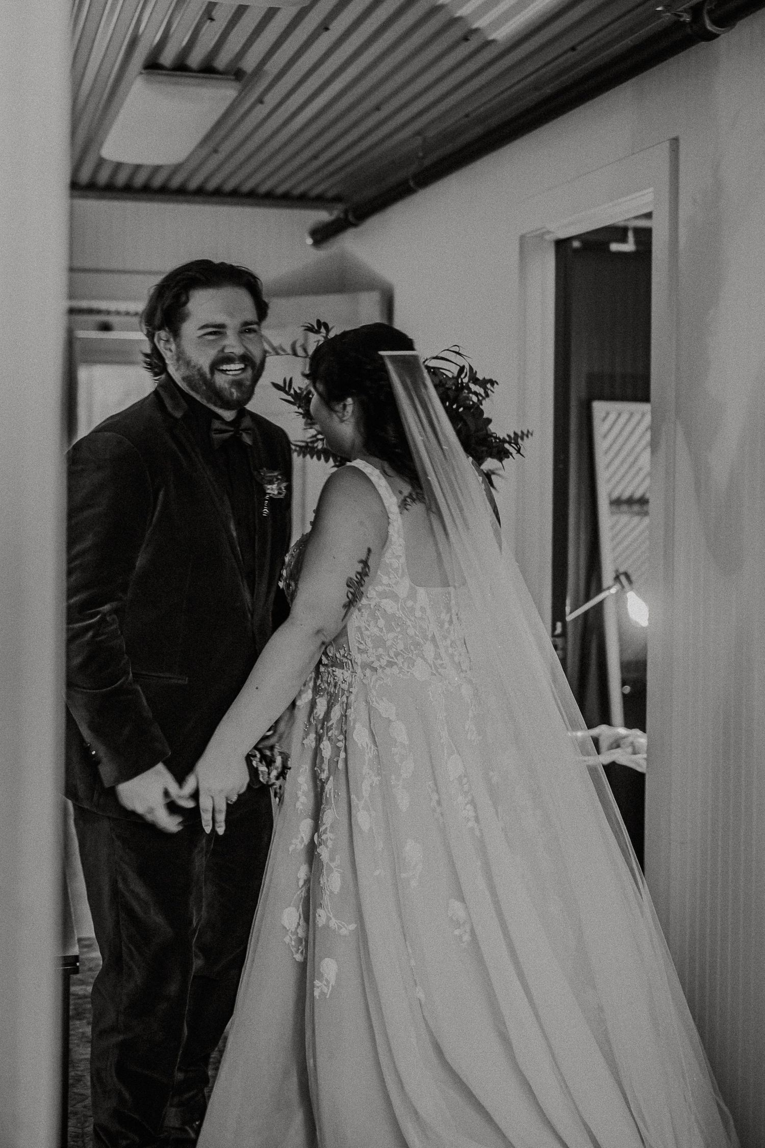 Black and white photo of groom smiling at bride in lace wedding gown during first look moment.