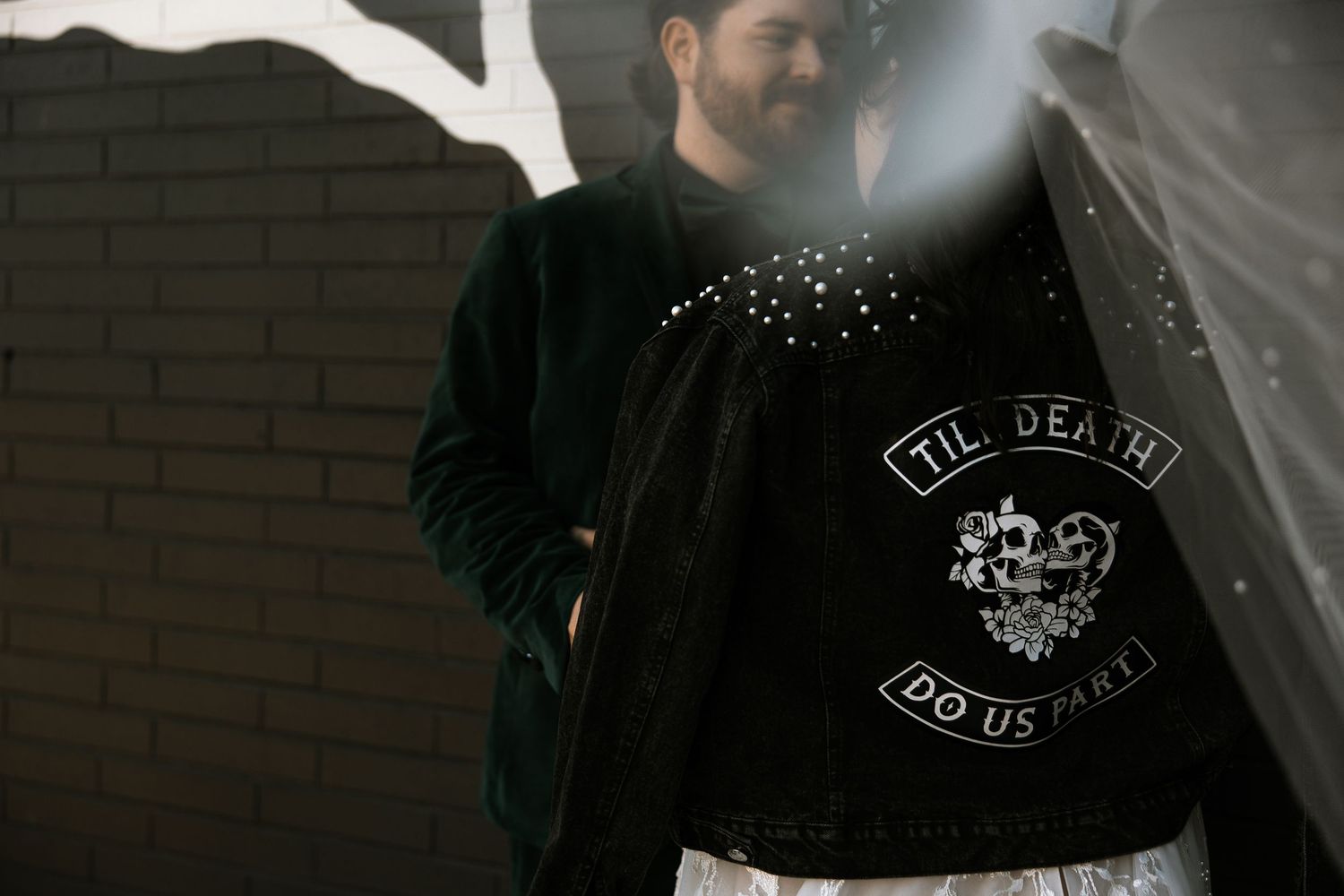Groom in black velvet jacket with Till Death Do Us Part embroidered patch smiles outdoors.