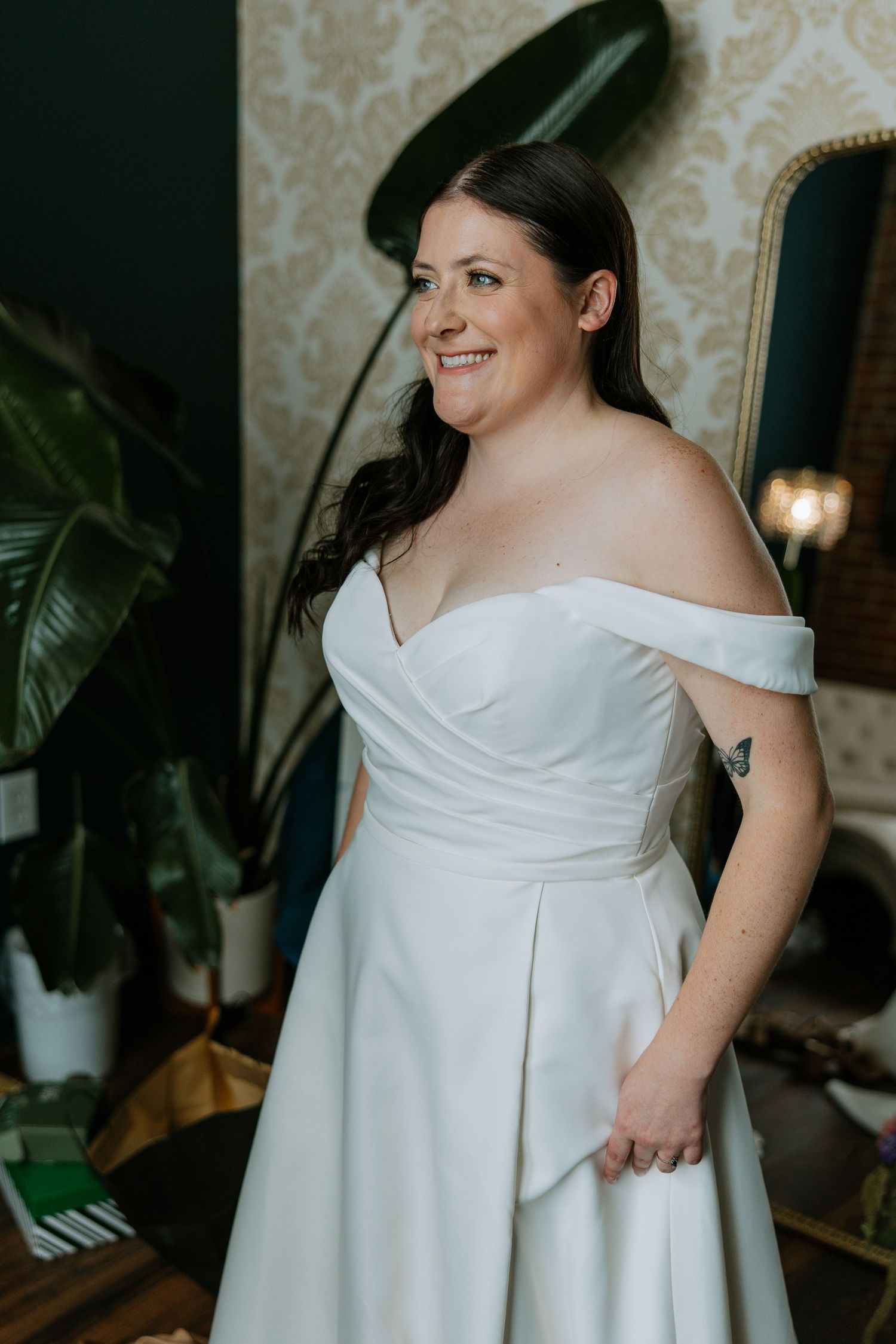 Smiling bride in off-shoulder white wedding gown posing in decorated bridal suite.
