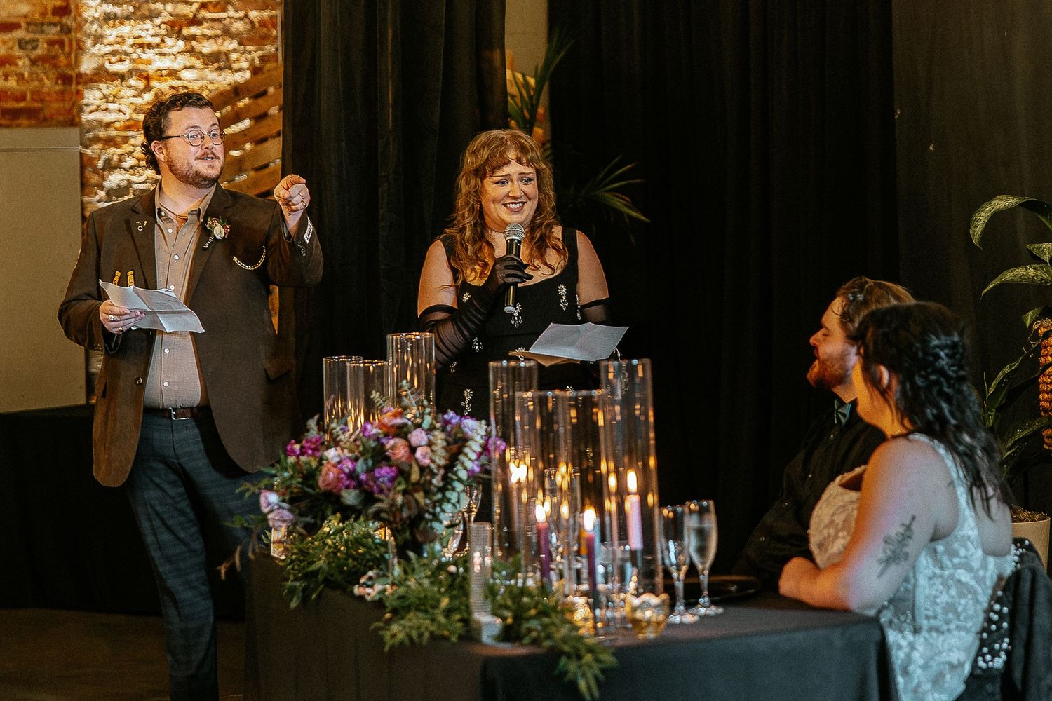 Two speakers deliver a heartfelt wedding toast at a candlelit reception table with guests listening.