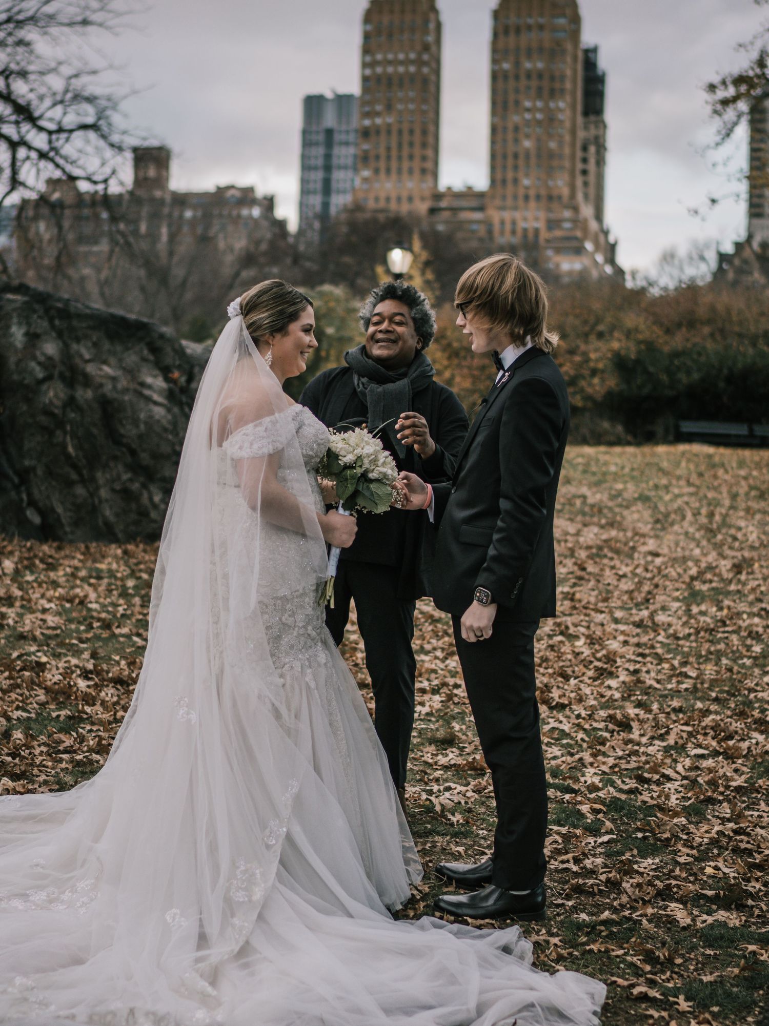 Joyful officiant celebrates with bride and groom after Central Park wedding ceremony among fall foliage.