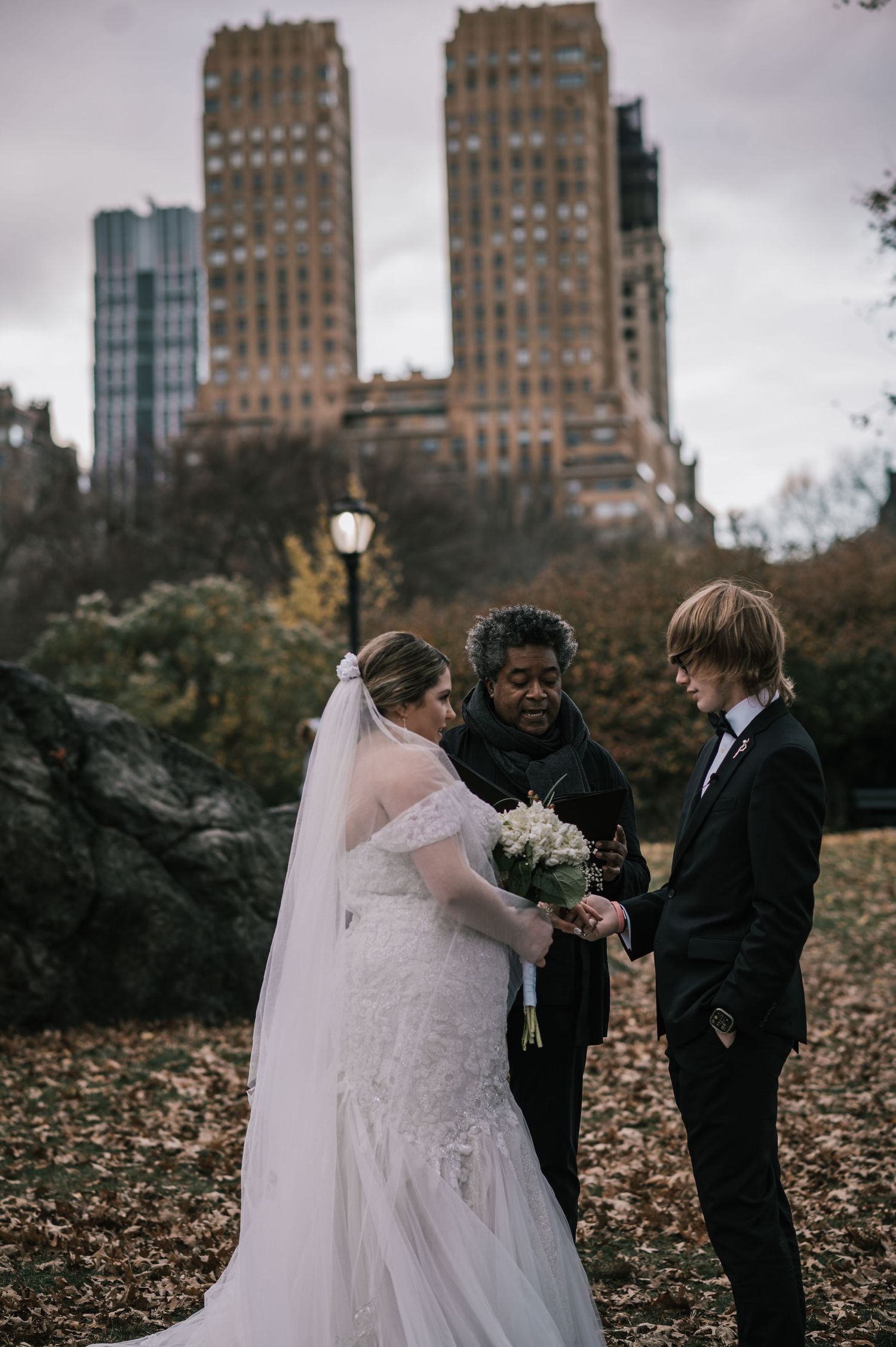 Bride and groom exchange vows with officiant in Central Park, NYC, surrounded by autumn leaves and city skyline.