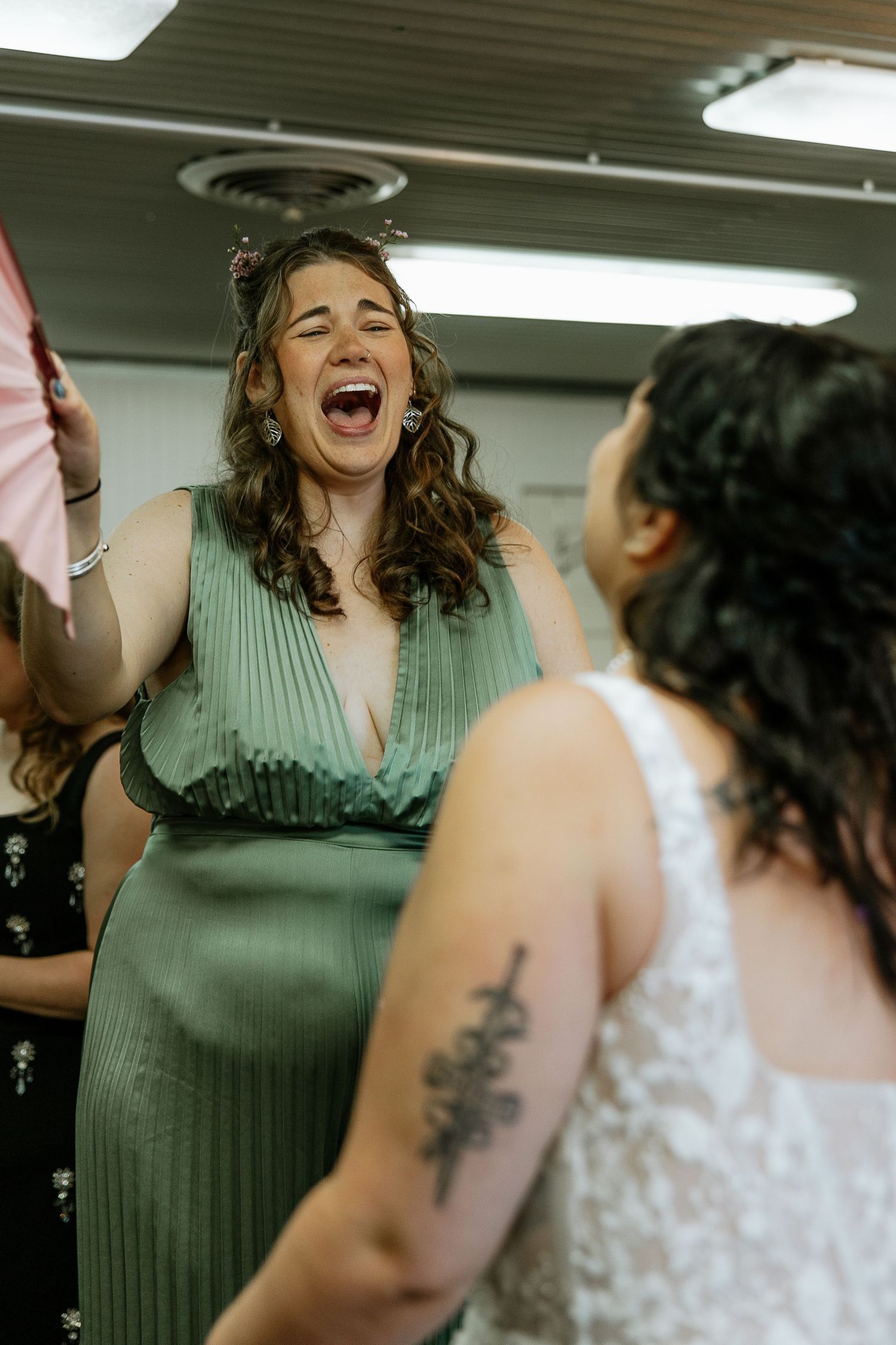Emotional bridesmaid in green dress laughing with bride in white lace gown before wedding.