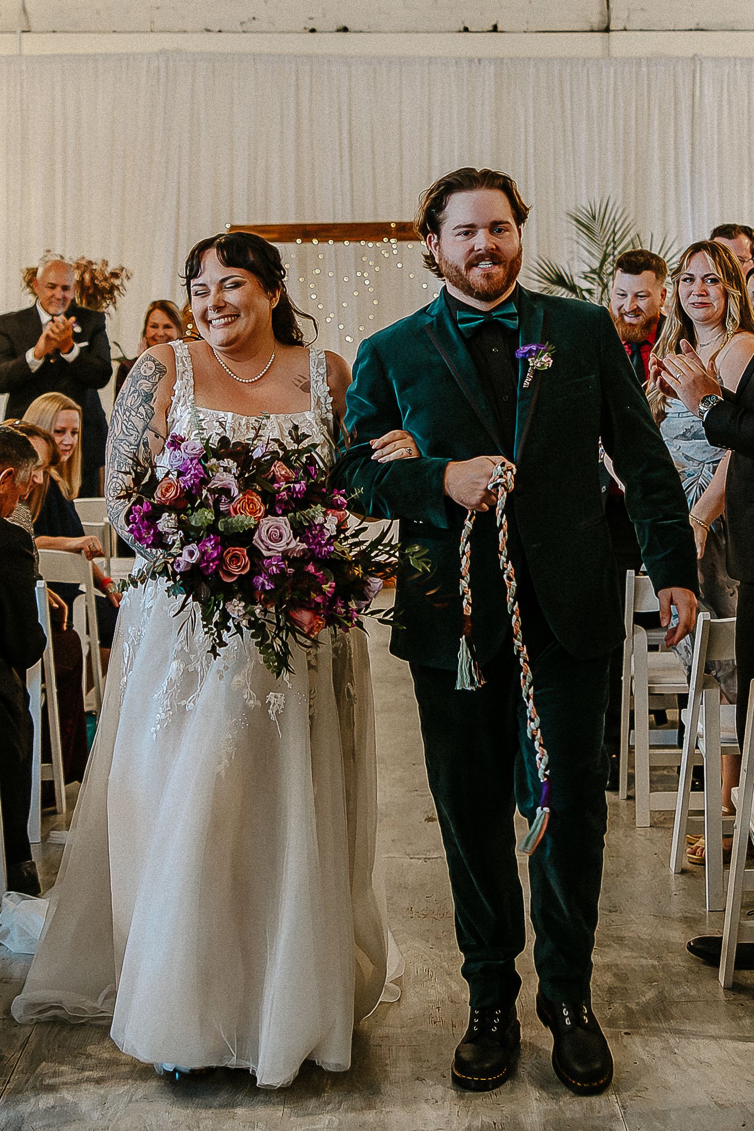 Smiling bride and groom walk down aisle, bride holding lush purple floral bouquet after ceremony.