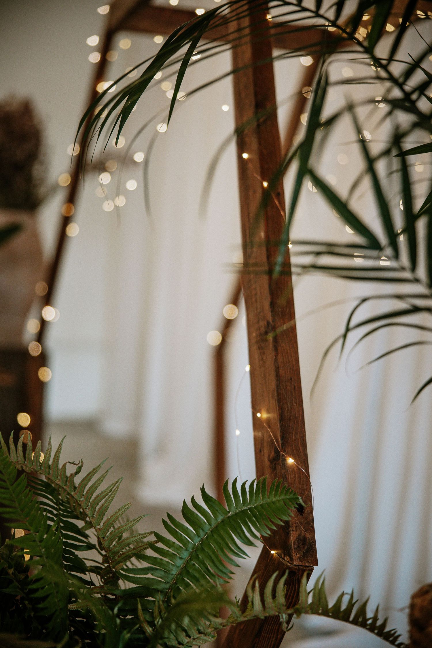 Close-up of wooden wedding arch with fairy lights bokeh and tropical fern greenery in foreground.